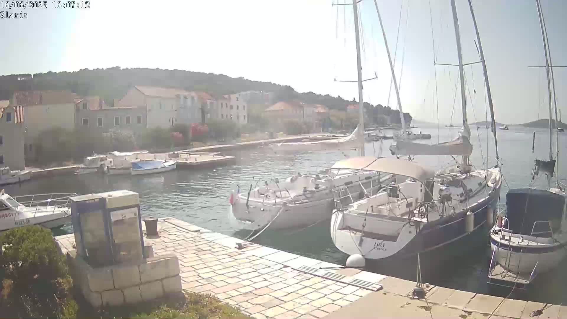 Several sailboats are moored in a harbor next to a stone quay, under a sunny sky.