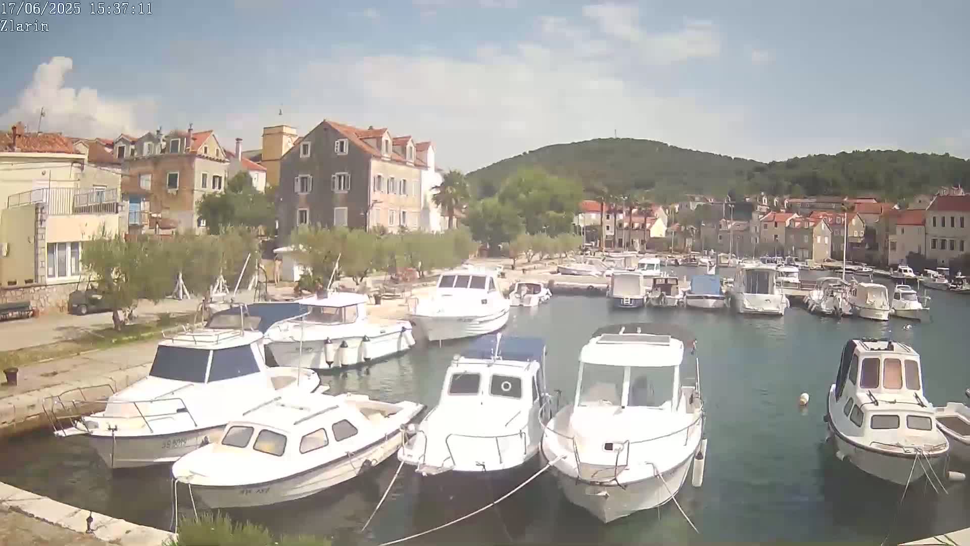 A harbor filled with numerous white motorboats is nestled beside a village of stone buildings under a mostly sunny sky.