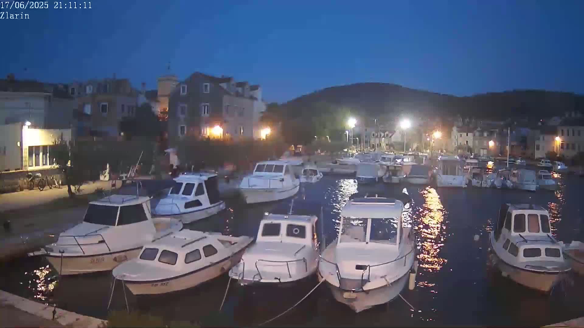 A harbor at night, filled with small motorboats, is situated next to a village under a dark blue twilight sky.