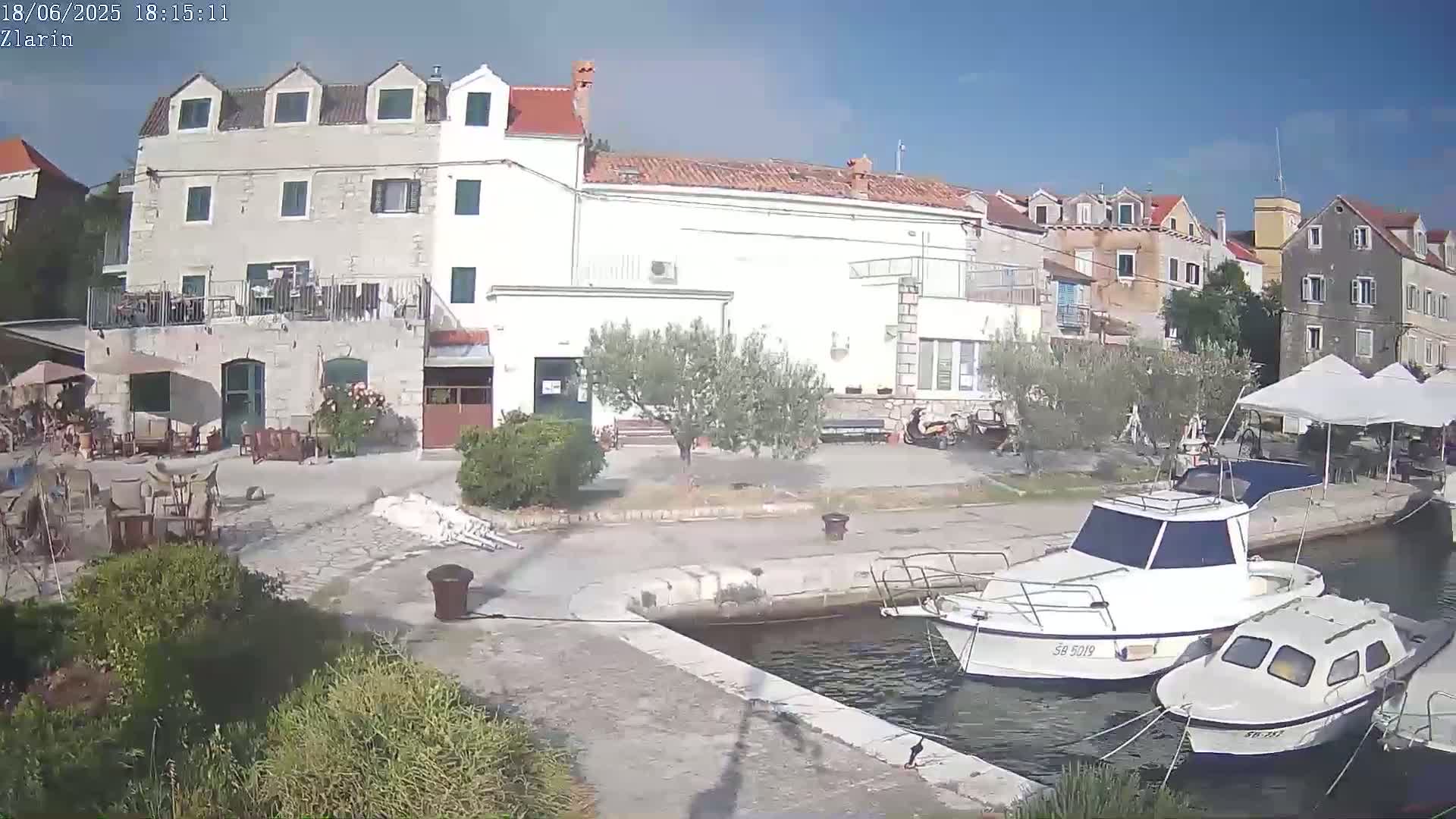 A sunny day reveals a waterfront scene with several buildings, outdoor seating, and two docked motorboats.