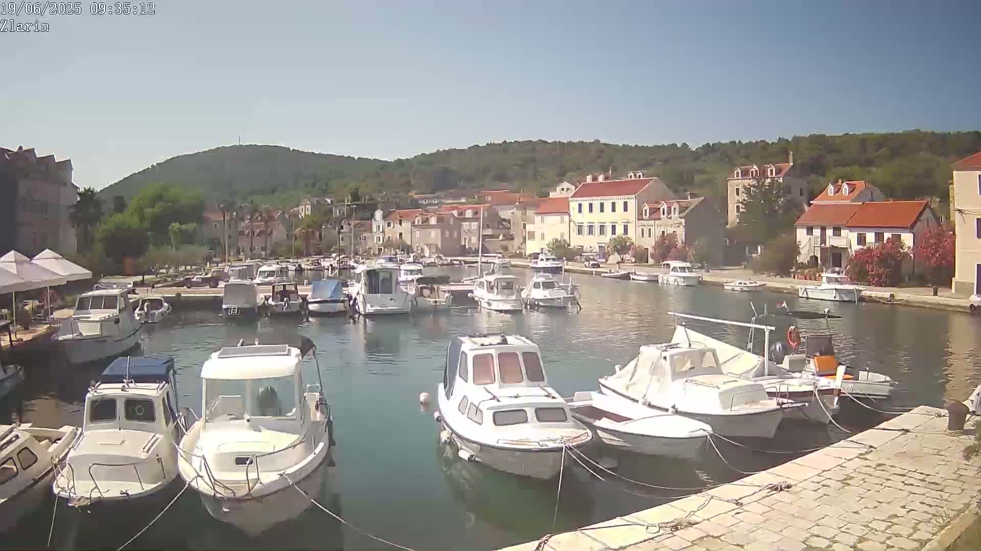A calm harbor filled with numerous boats is nestled beside a village of low-rise buildings under a clear, sunny sky.