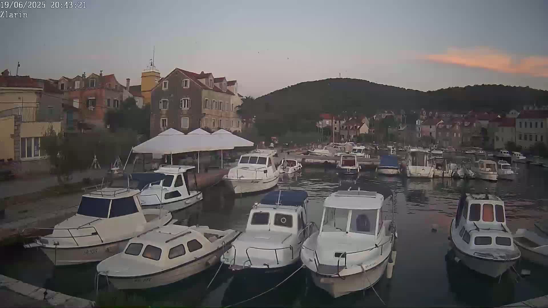 Many small motorboats are docked in a calm harbor next to a village of stone buildings at dusk under a mostly clear sky.
