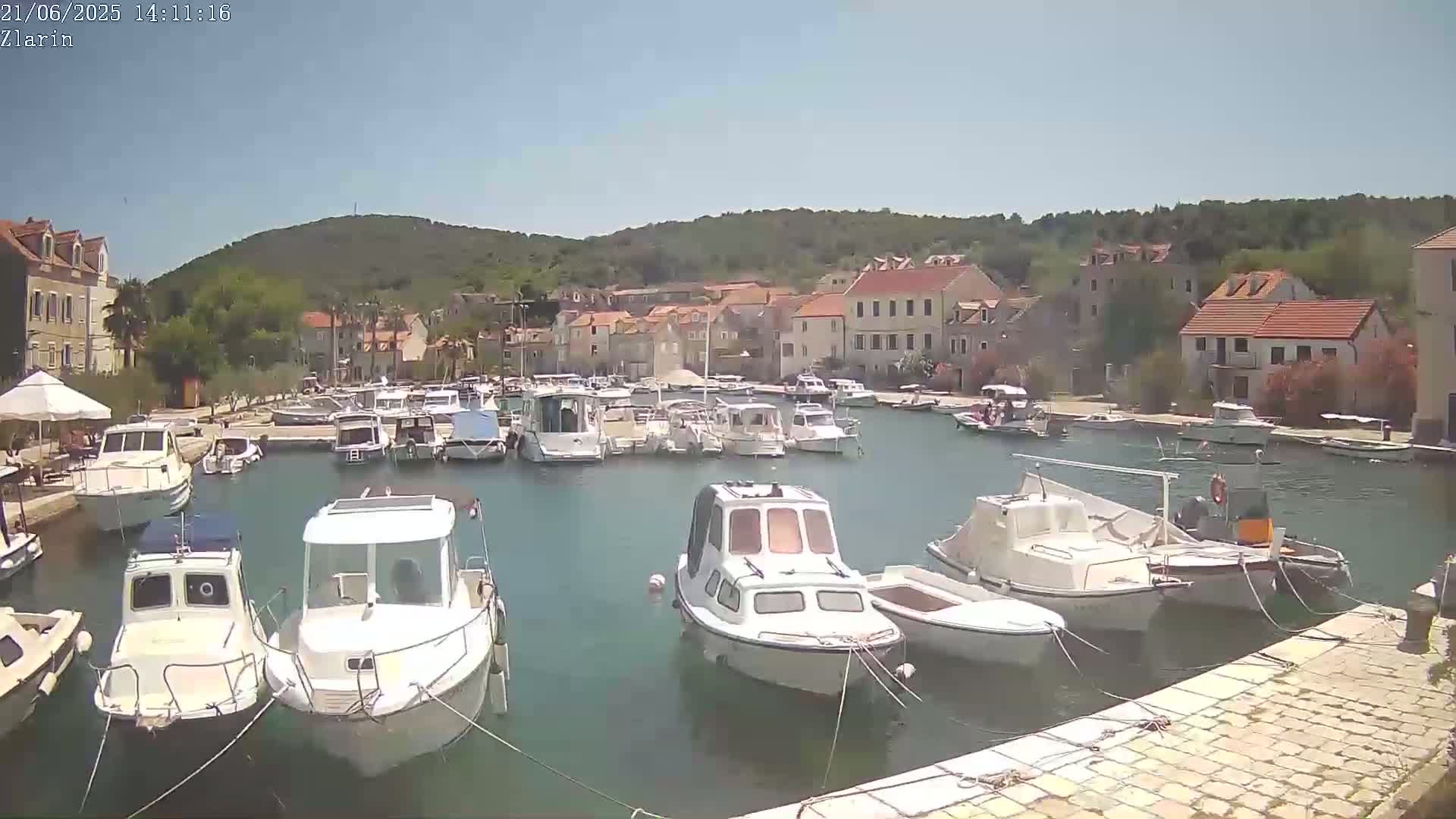 A harbor filled with numerous boats is nestled in a small town under a clear, sunny sky, backed by a verdant hill.