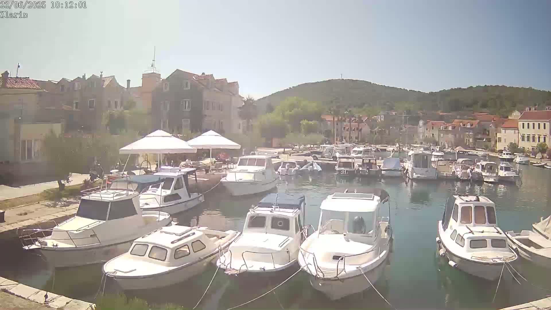A harbor filled with numerous white motorboats is nestled beside a waterfront town on a sunny day, with a verdant hill rising in the background.