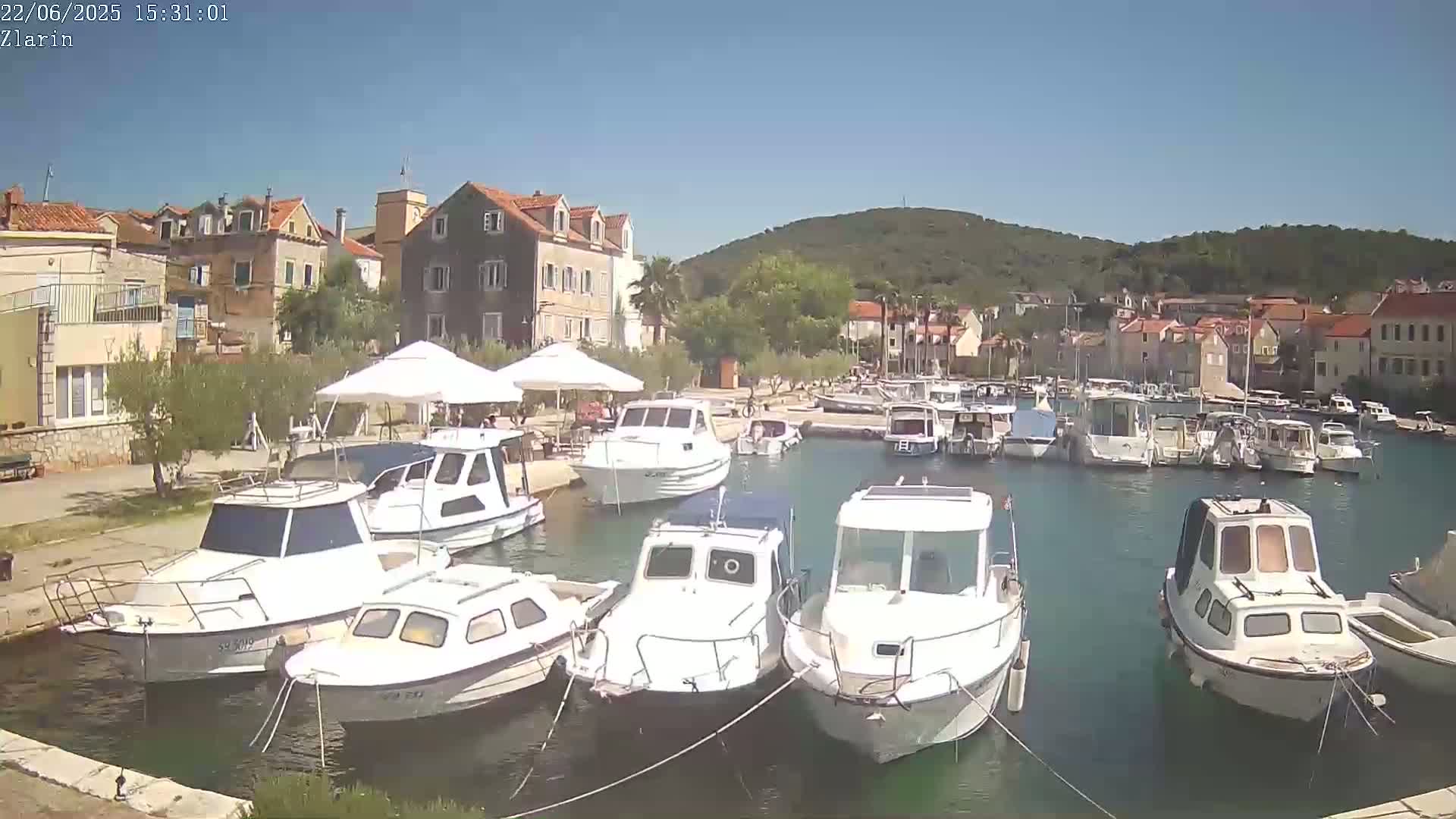 A sunny harbor scene shows numerous white motorboats moored in calm, clear water, alongside a waterfront lined with buildings and shaded outdoor seating areas.