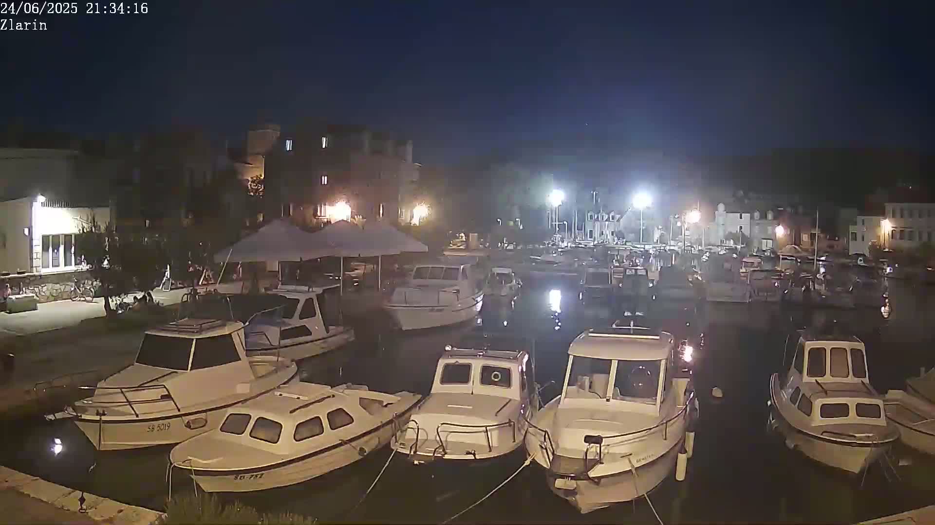 A nighttime harbor scene shows numerous small motorboats moored in calm water, illuminated by artificial lights against a dark sky.