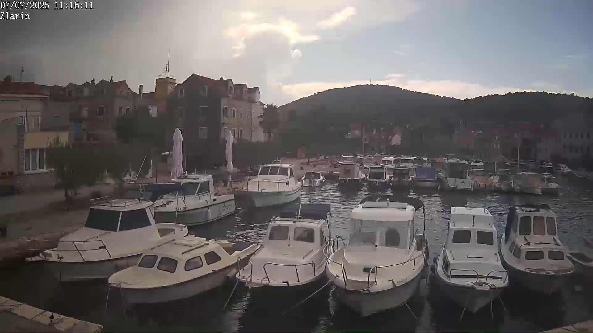 Many small motorboats are moored in a calm harbor next to a village of stone buildings under a partly cloudy sky.