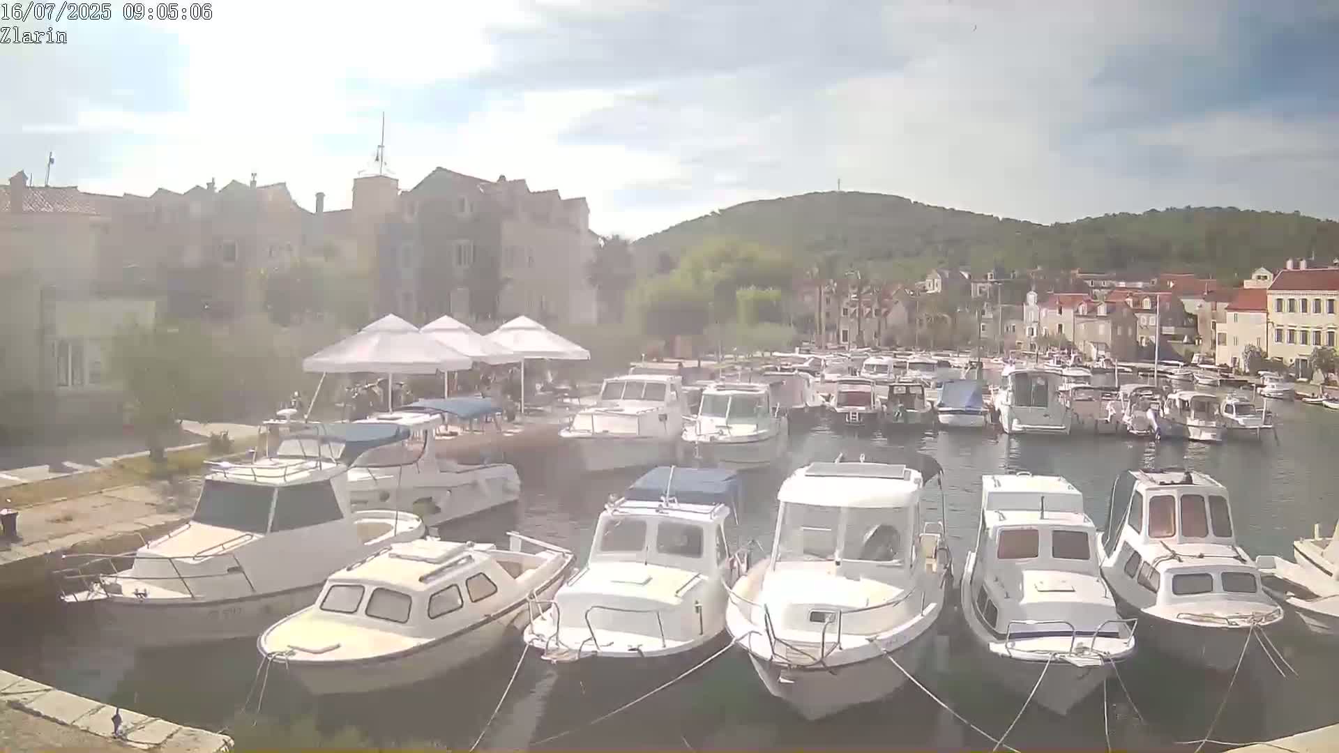 Many white motorboats are docked in a harbor next to a town on a partly cloudy day.