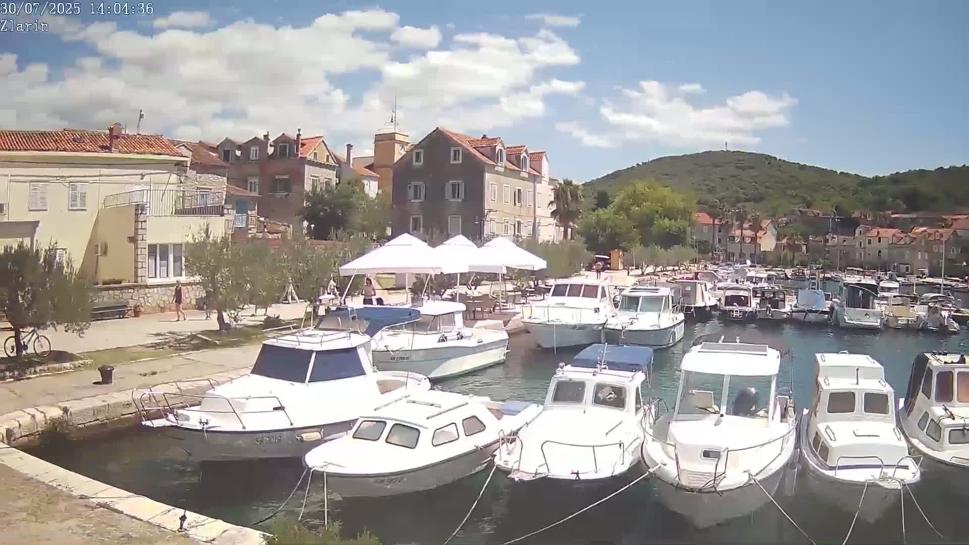 A hazy sunny day reveals a waterfront scene with buildings, boats docked at a quay, and outdoor seating areas under umbrellas.