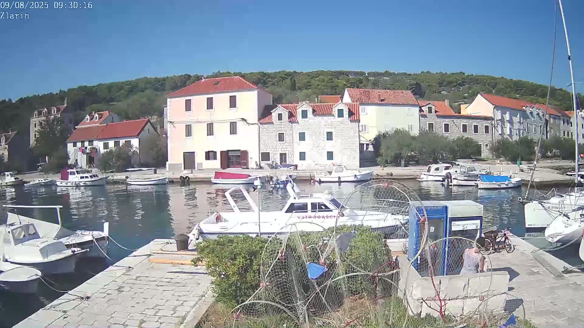 A sunny day shows a calm harbor filled with boats, alongside a waterfront village of stone and stucco buildings, backed by green hills.