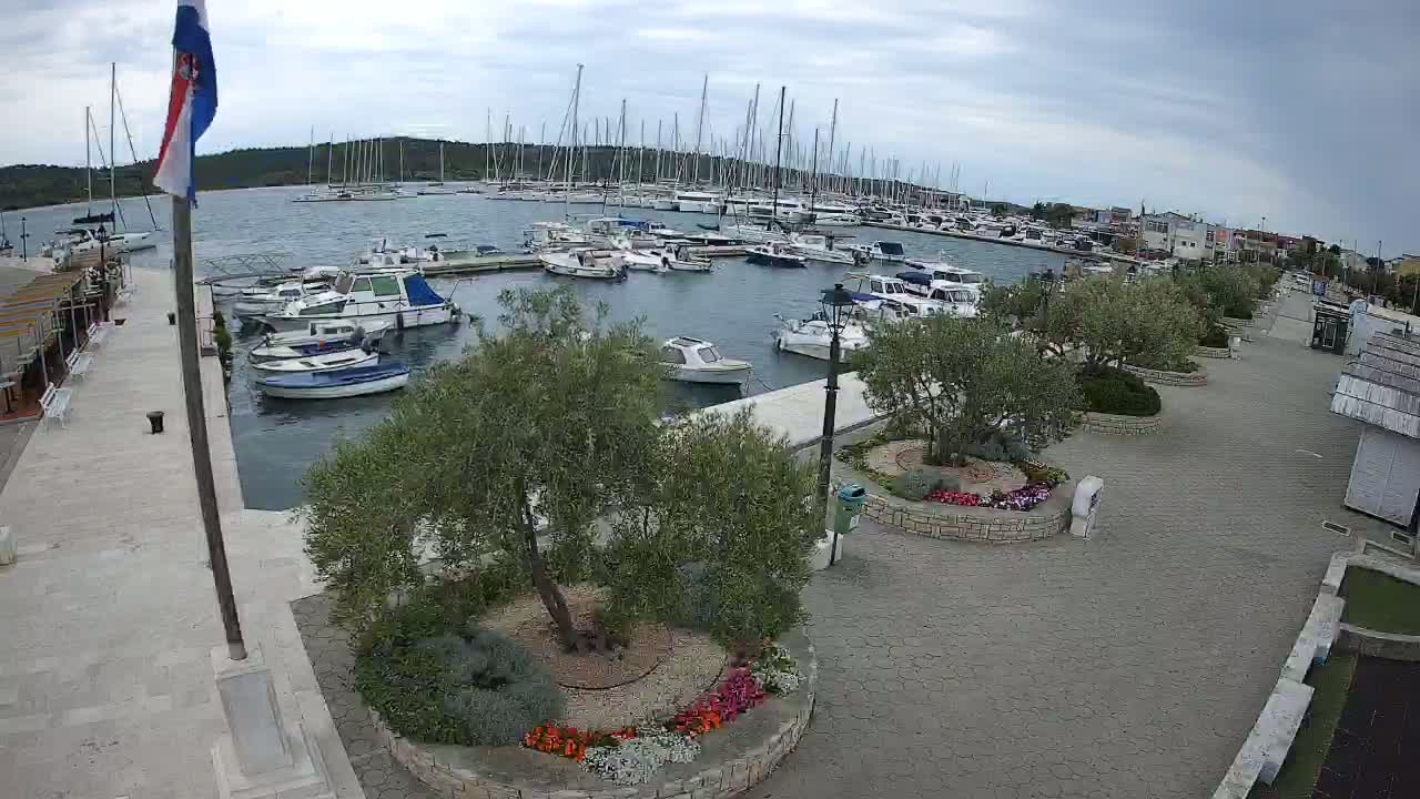 A marina filled with sailboats and motorboats is viewed from above on a partly cloudy day, with a paved walkway and landscaped areas nearby.