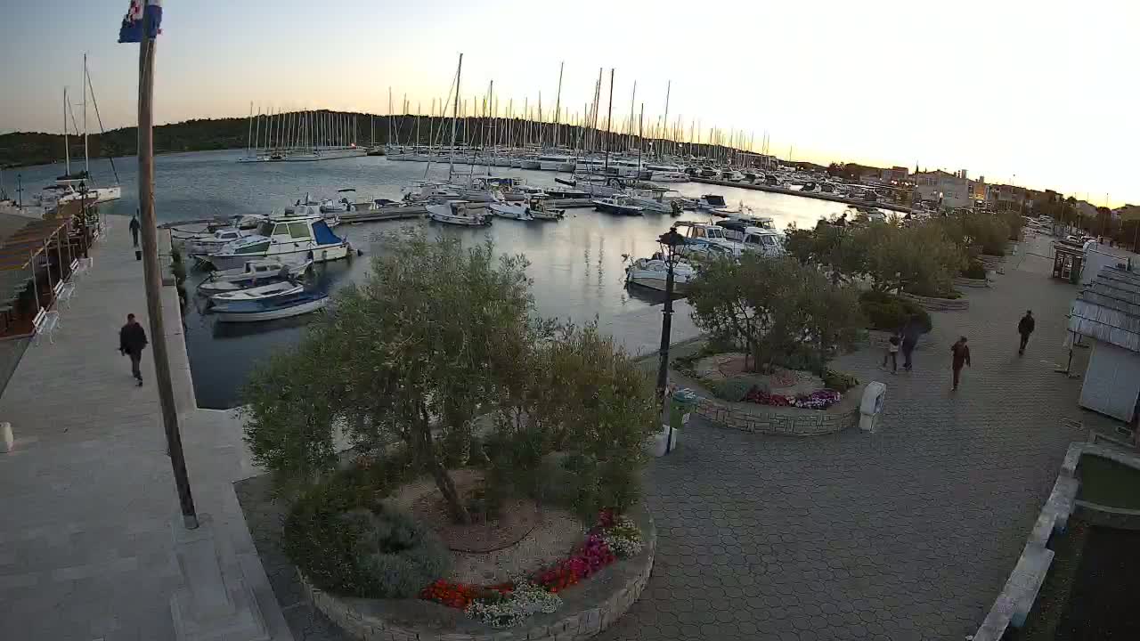A marina filled with sailboats and motorboats at sunset, with people walking along a paved walkway lined with landscaping.