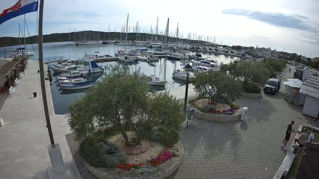 A marina filled with sailboats and motorboats on a partly cloudy day, next to a paved waterfront area with landscaping and a few people walking by.