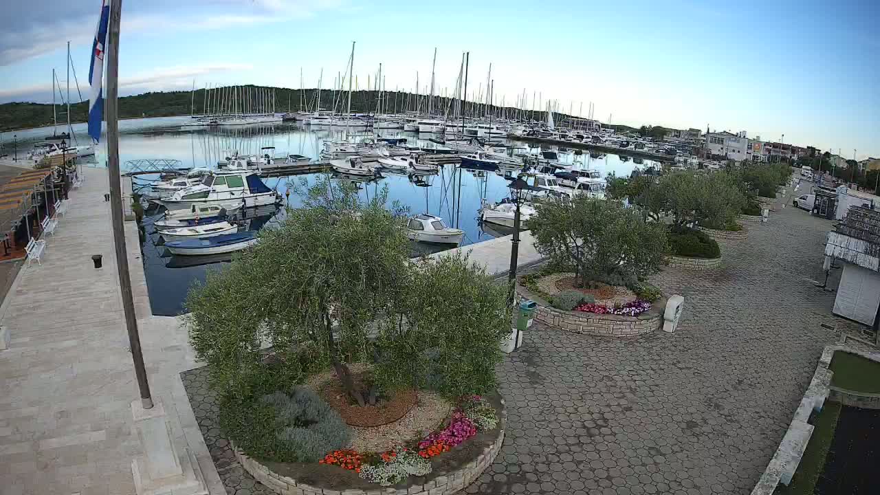 A marina filled with sailboats and motorboats is viewed from above on a partly cloudy day, with a paved walkway and landscaped areas visible in the foreground.