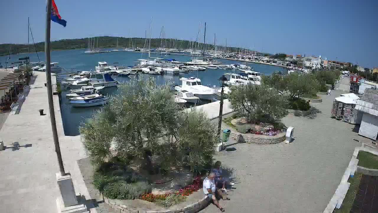 A marina filled with boats is shown on a sunny day, with a paved waterfront area featuring landscaping and two people sitting on a low wall.