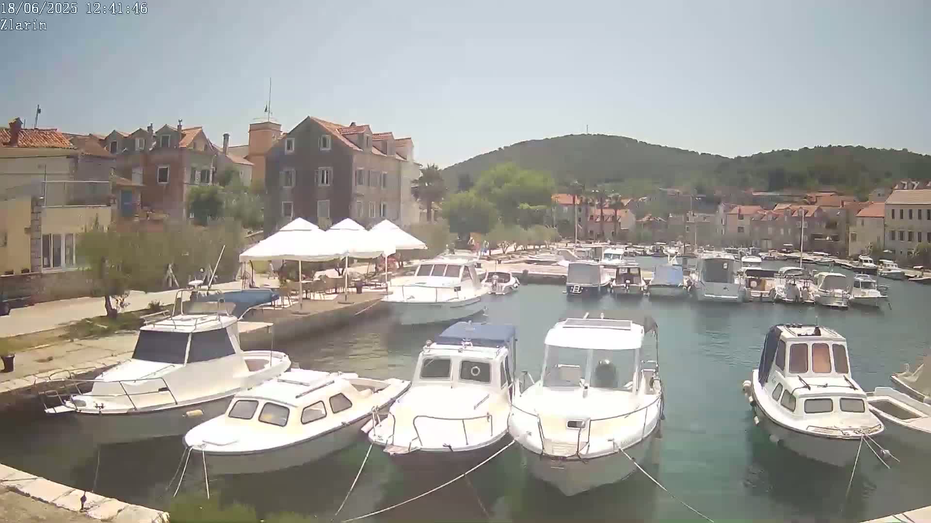 Several white motorboats are moored in a calm harbor next to a village of low-rise buildings on a sunny day.