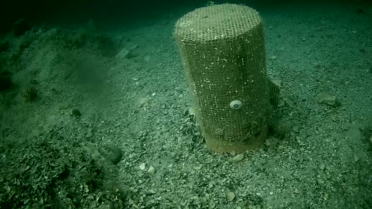 An underwater scene shows a cylindrical object encased in mesh netting resting on a dark, gravelly seabed.