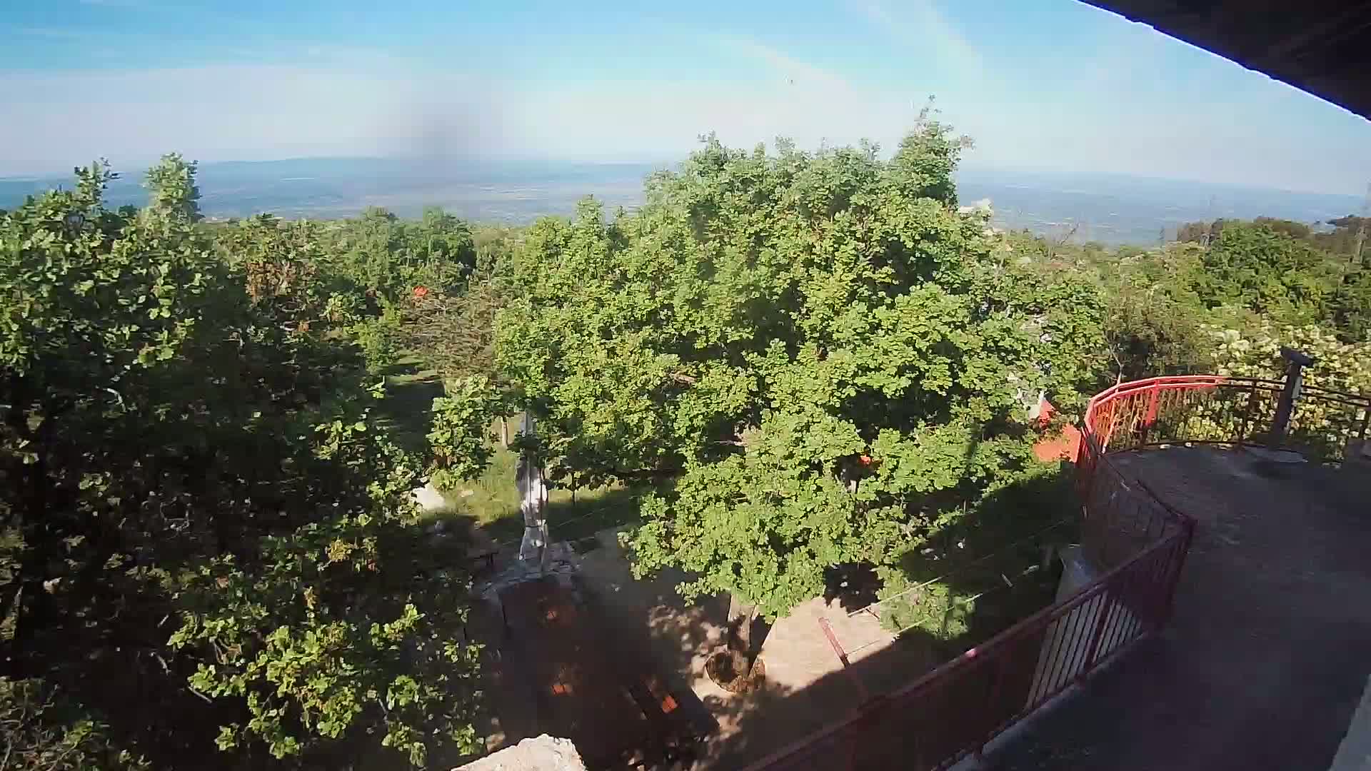 A high-angle view shows a leafy, green landscape with picnic tables below a partially visible balcony railing on a sunny day, overlooking a distant body of water.