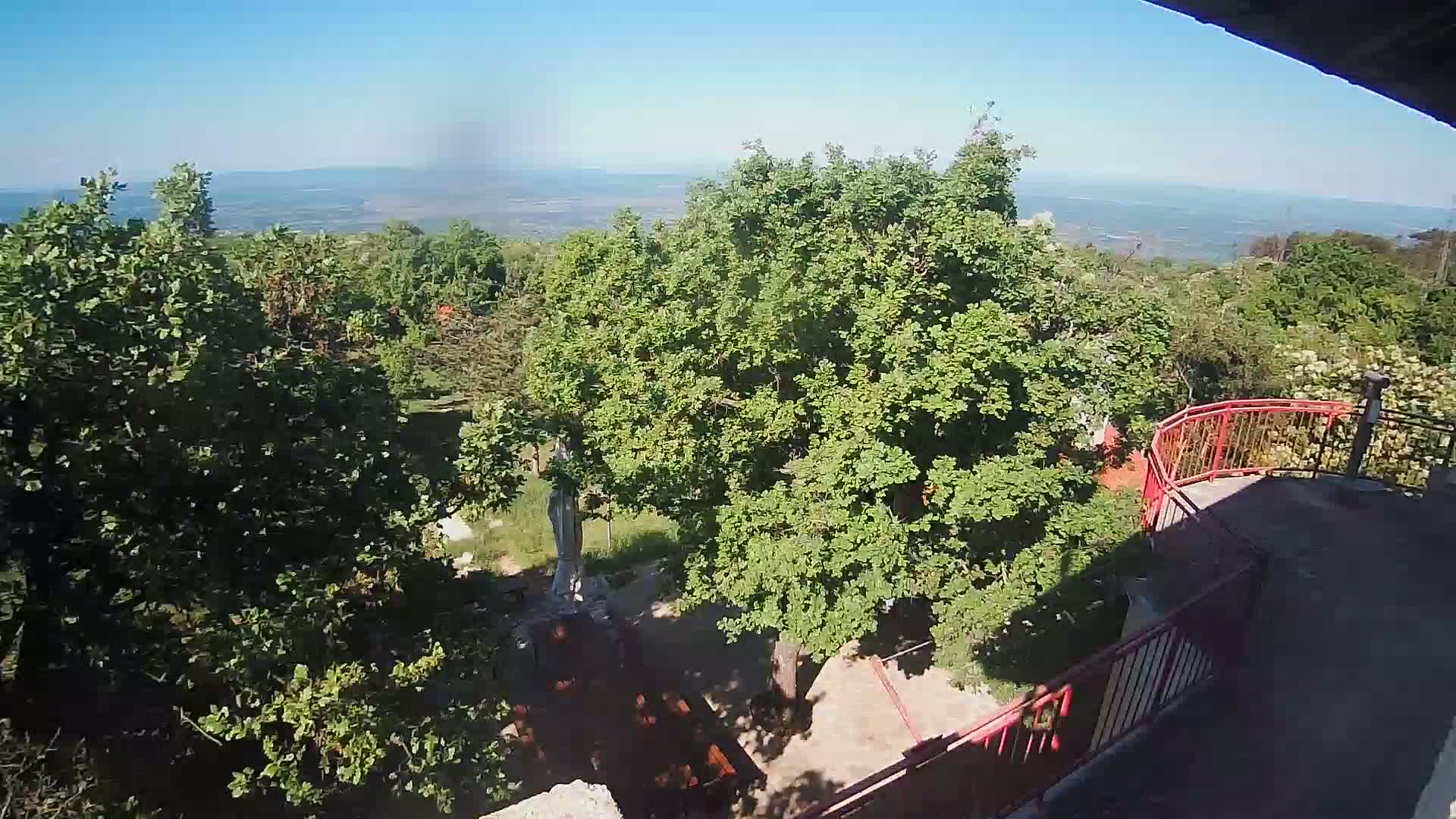 A high-angle view shows a statue among lush green trees and a distant, hazy view of a plain under a clear blue sky.