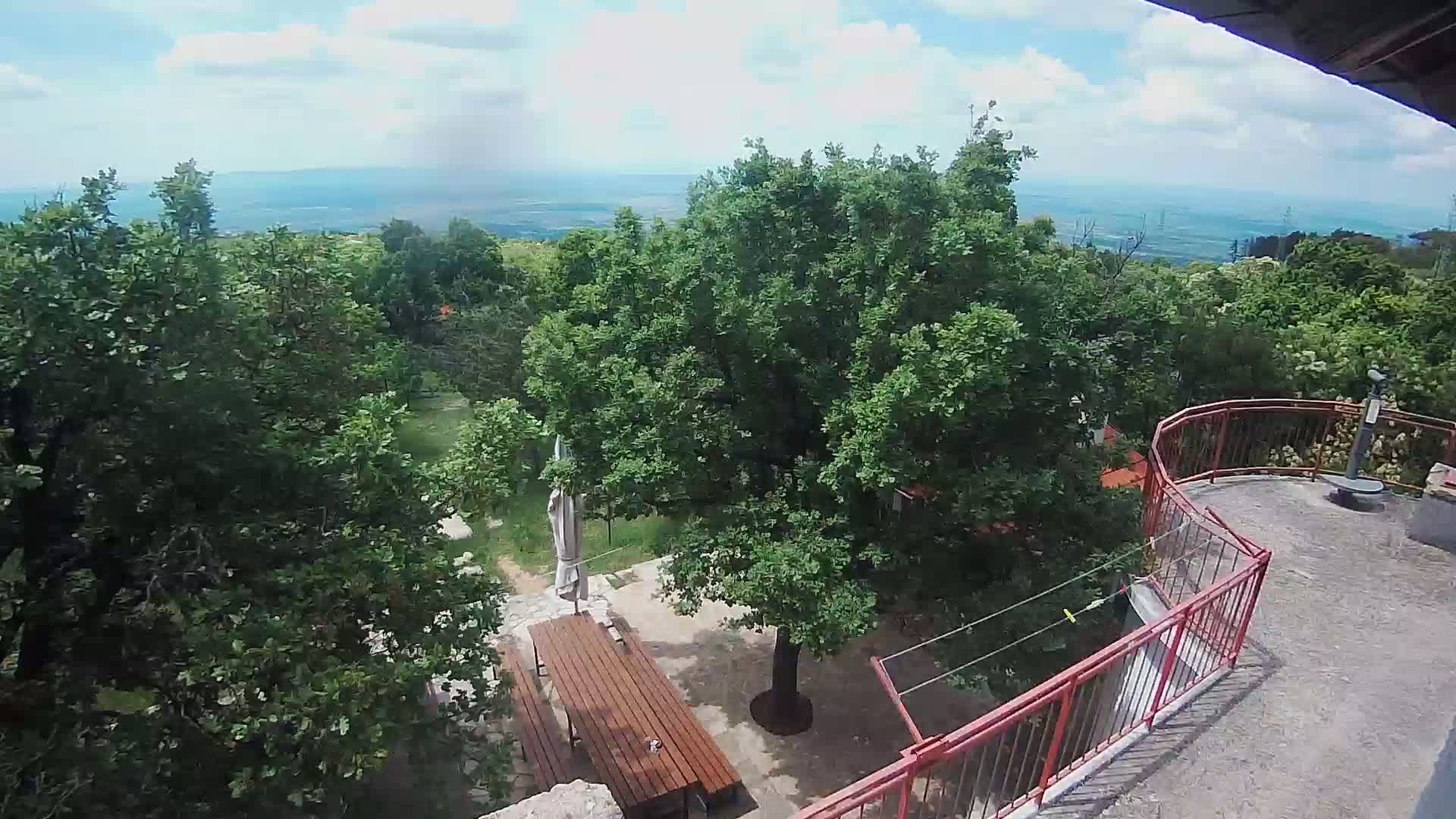 An elevated view shows a patio with a wooden table and trees, overlooking a sea under a partly cloudy sky.
