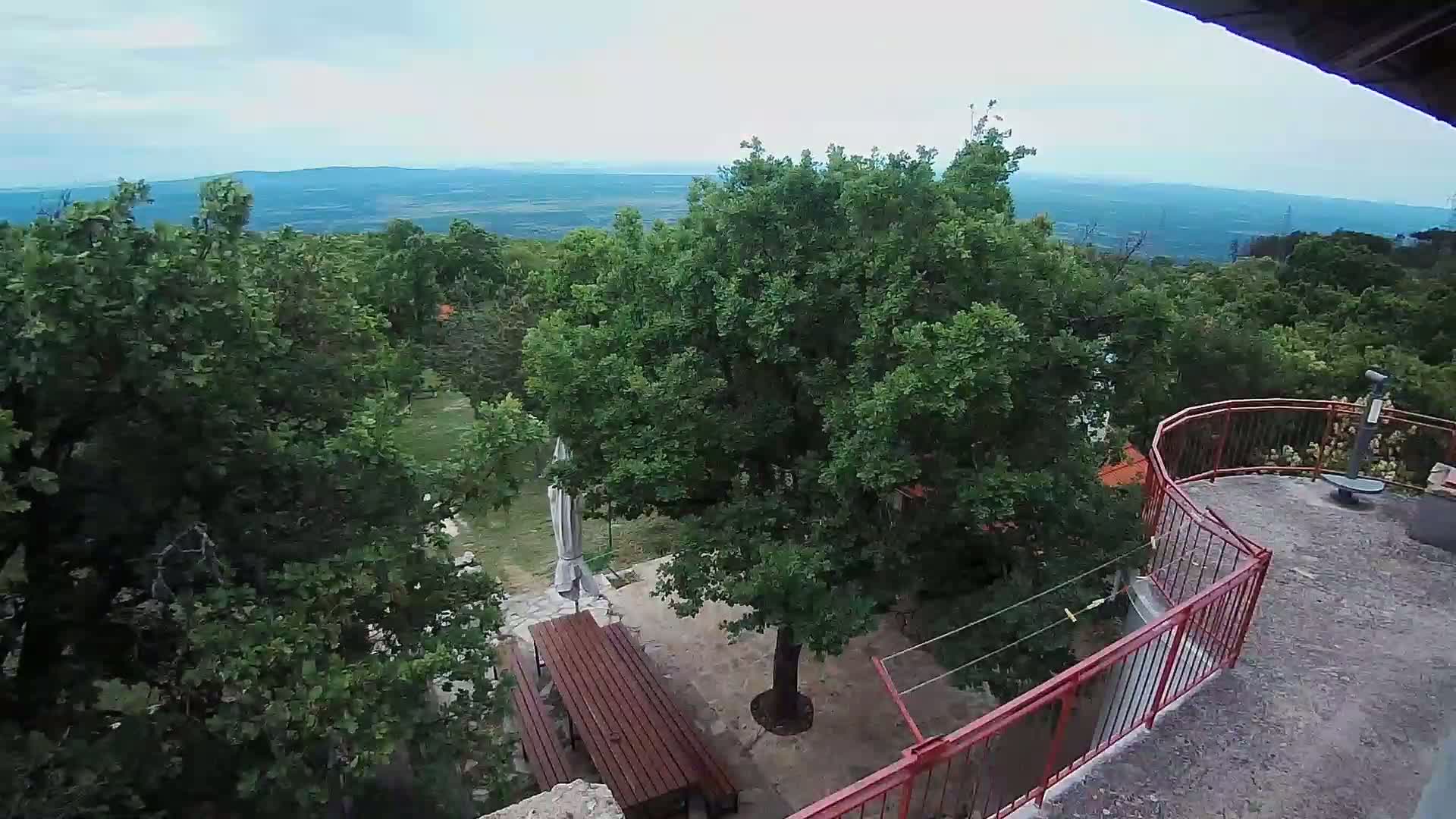 A patio with picnic tables is nestled among leafy green trees on a hill overlooking a distant valley under a cloudy sky.