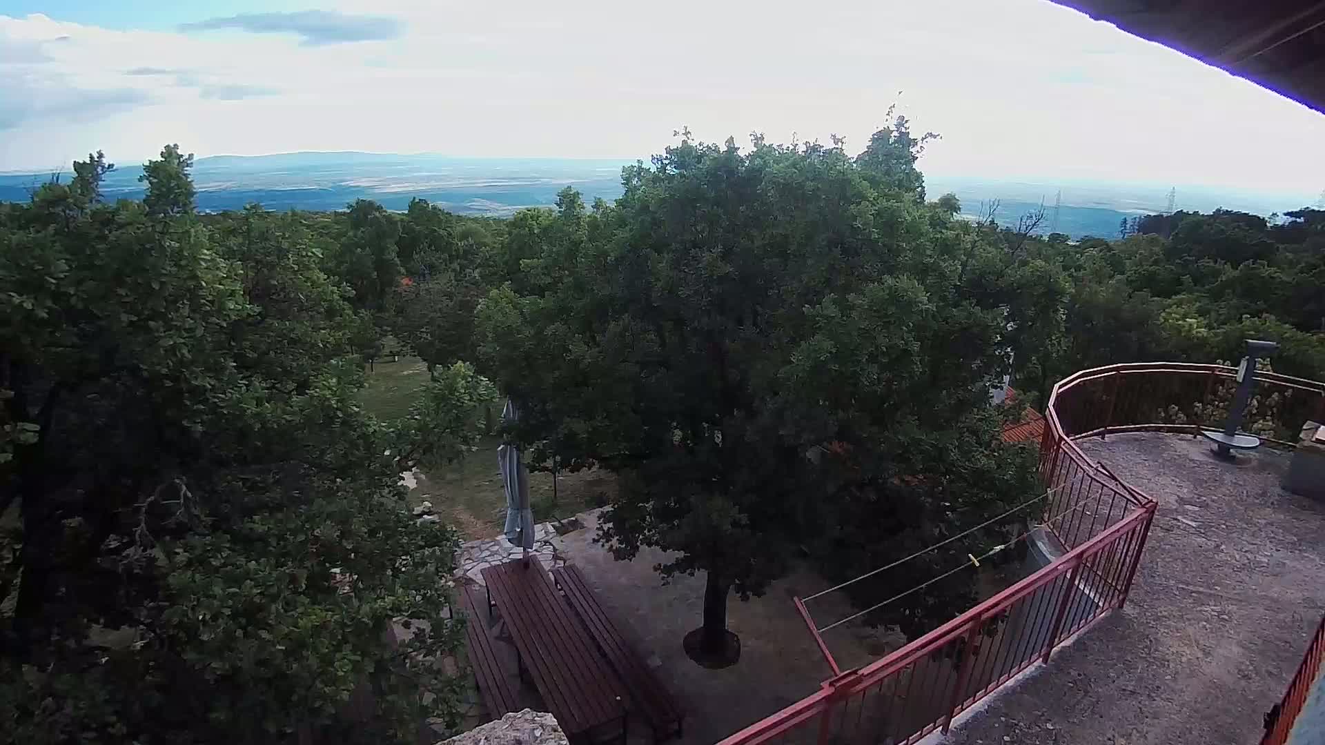 A partly cloudy day overlooks a patio with picnic tables beneath a large tree and a red railing overlooking a verdant landscape.