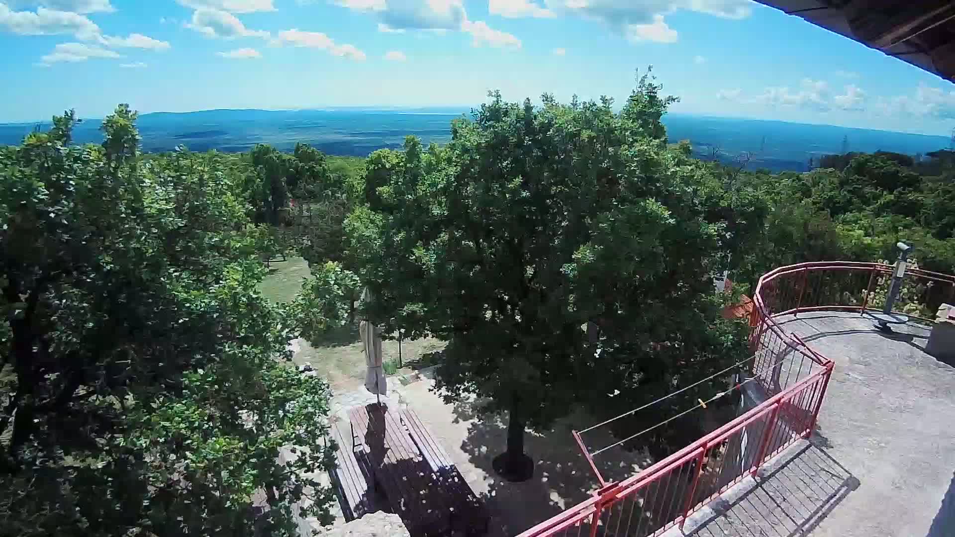 An elevated view shows a partly sunny day overlooking a lush green landscape with a distant mountain range, featuring a patio with a red railing and outdoor furniture situated amongst leafy trees.