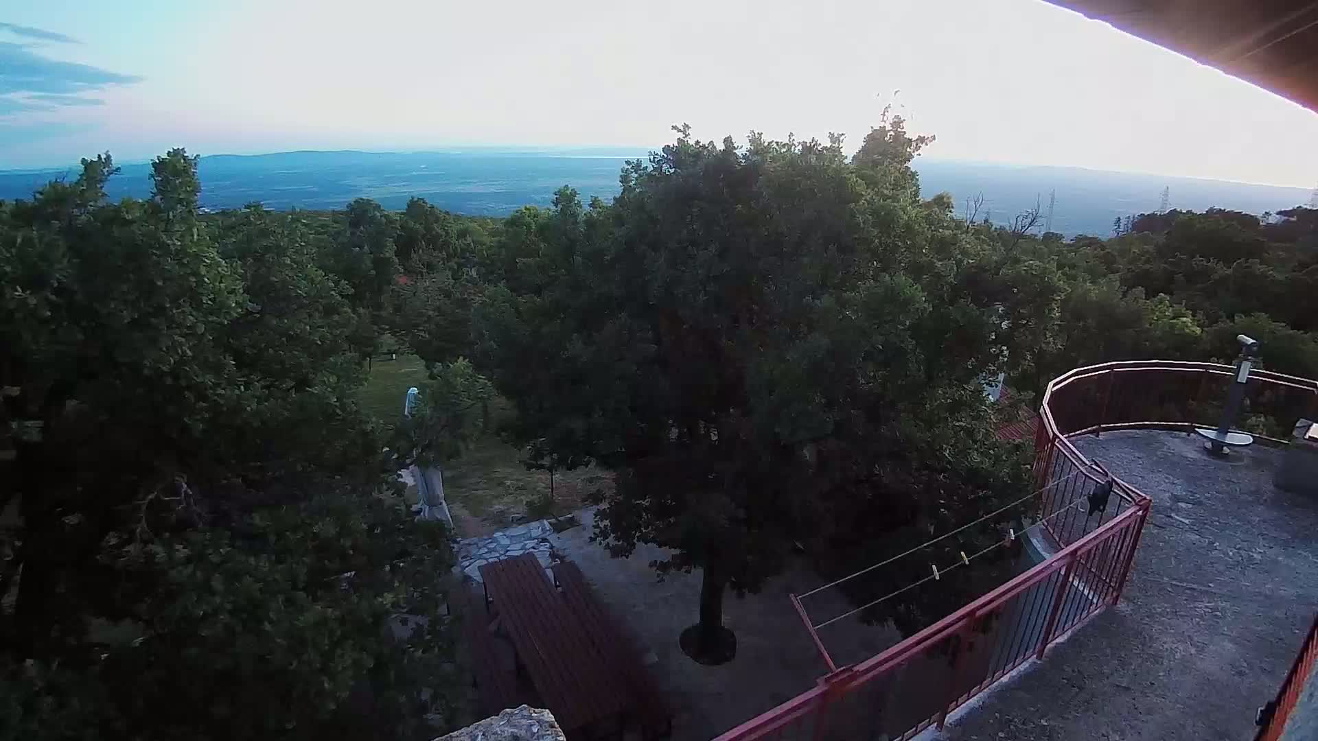 An elevated view shows a patio area with picnic tables surrounded by lush green trees, overlooking a distant valley under a clear, light-blue sky.