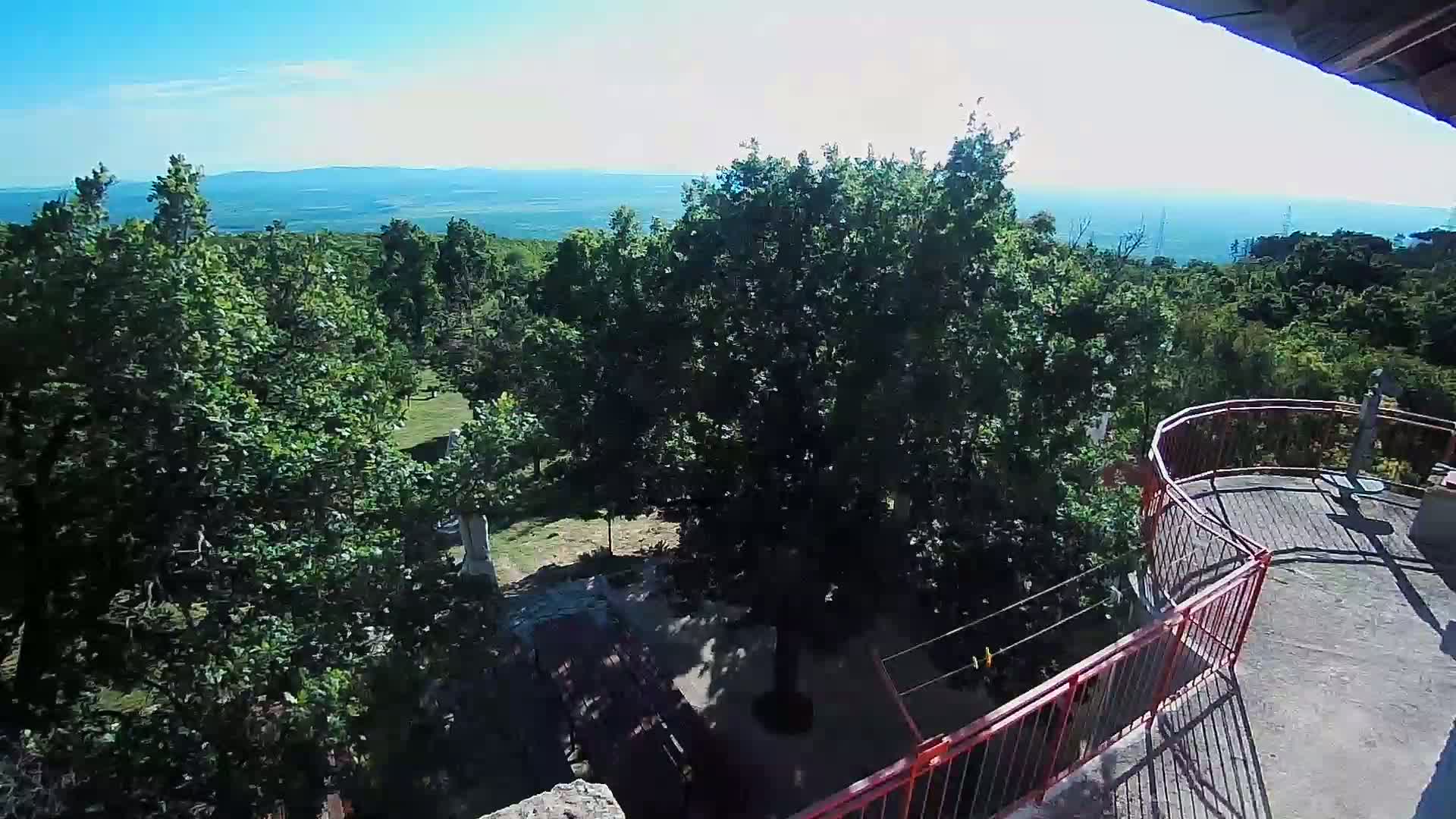 A high-angle view shows a lush, green landscape with a distant ocean under a clear, sunny sky, partially visible from a red-railed balcony.