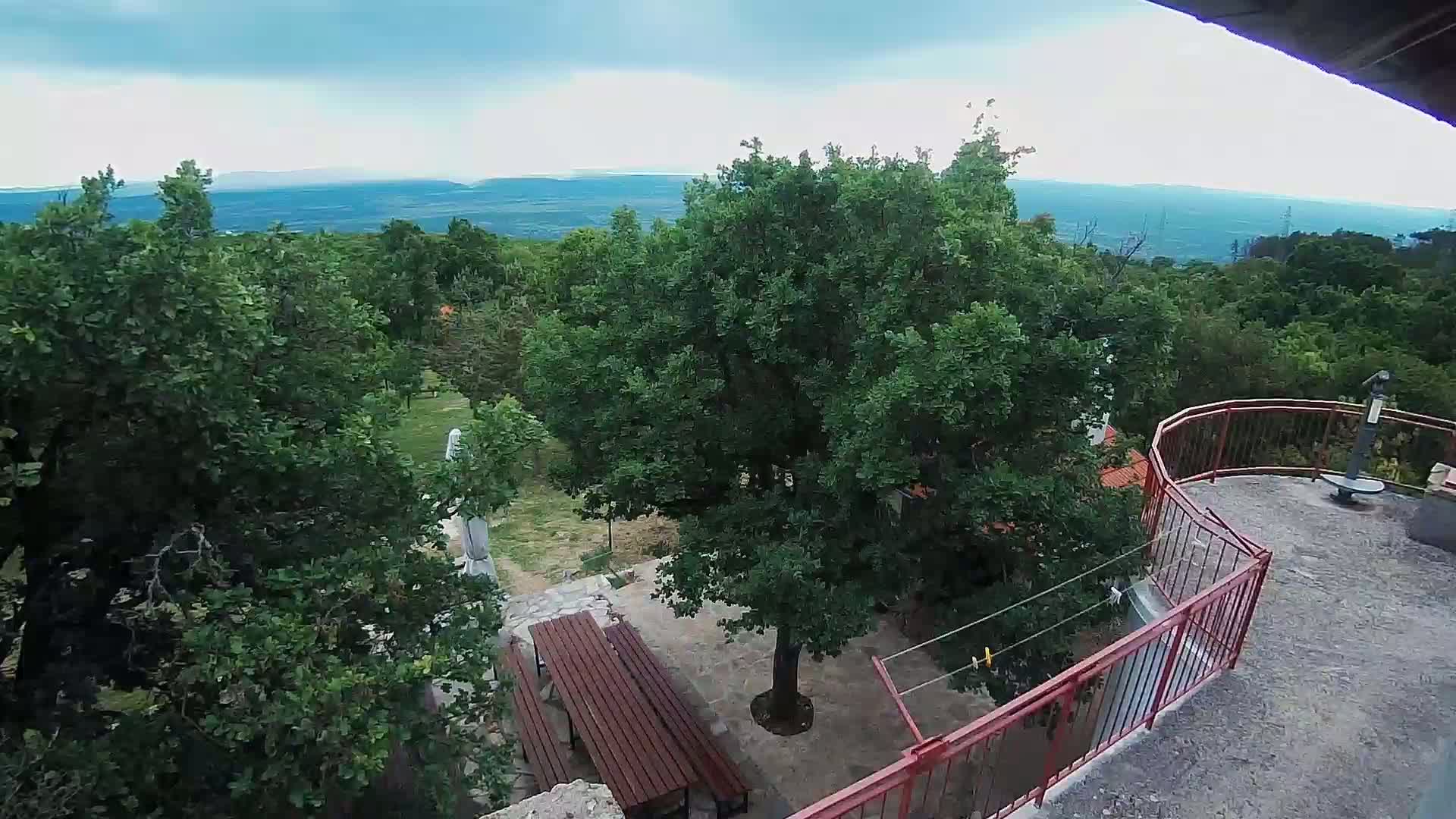 An outdoor patio with a picnic table is surrounded by lush green trees, overlooking a distant mountain range under a partly cloudy sky.