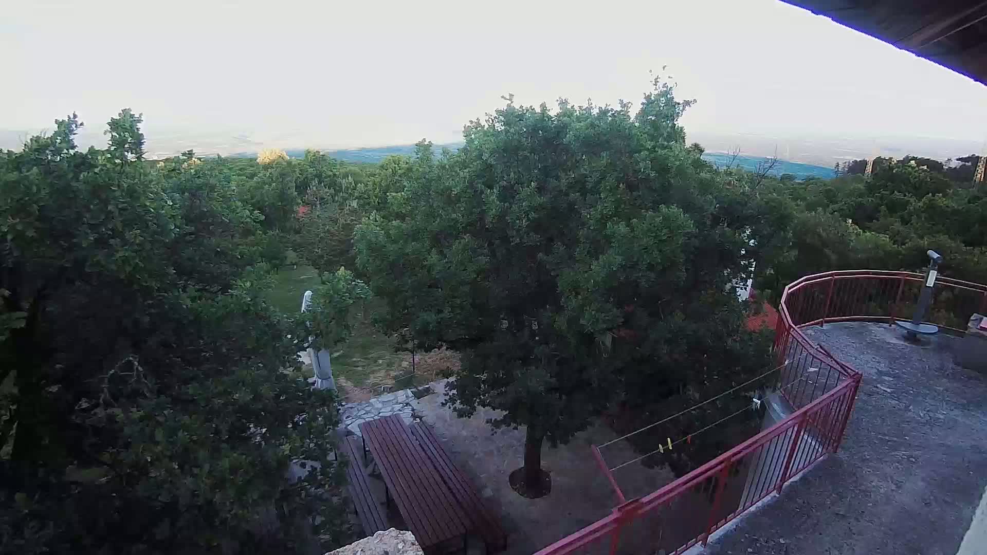 An elevated view shows a patio with a wooden table and chairs nestled among lush green trees on a partly cloudy day, overlooking a distant mountain range.