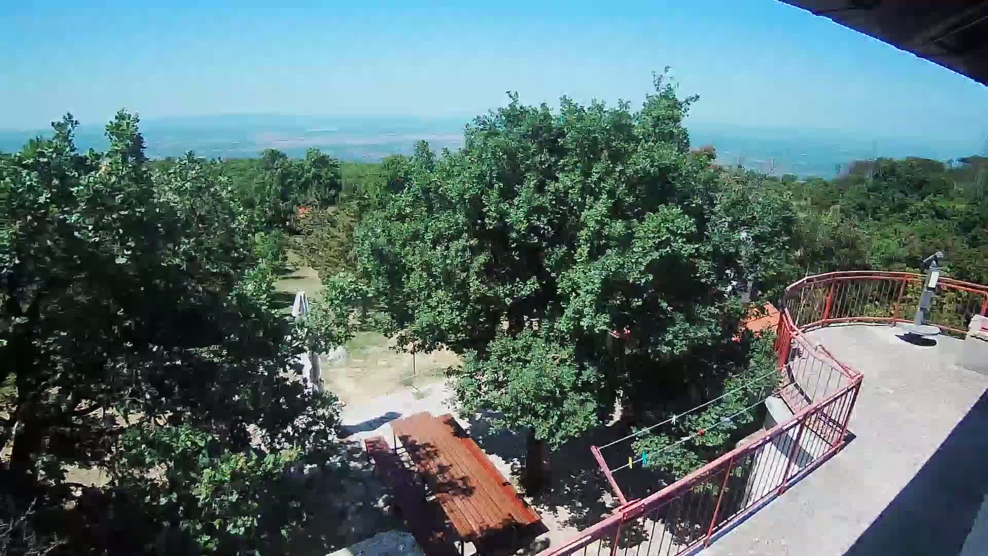 A high-angle view shows a sunny outdoor scene with lush green trees, a wooden picnic table, a red railing, and a distant view of a hazy plain under a clear blue sky.