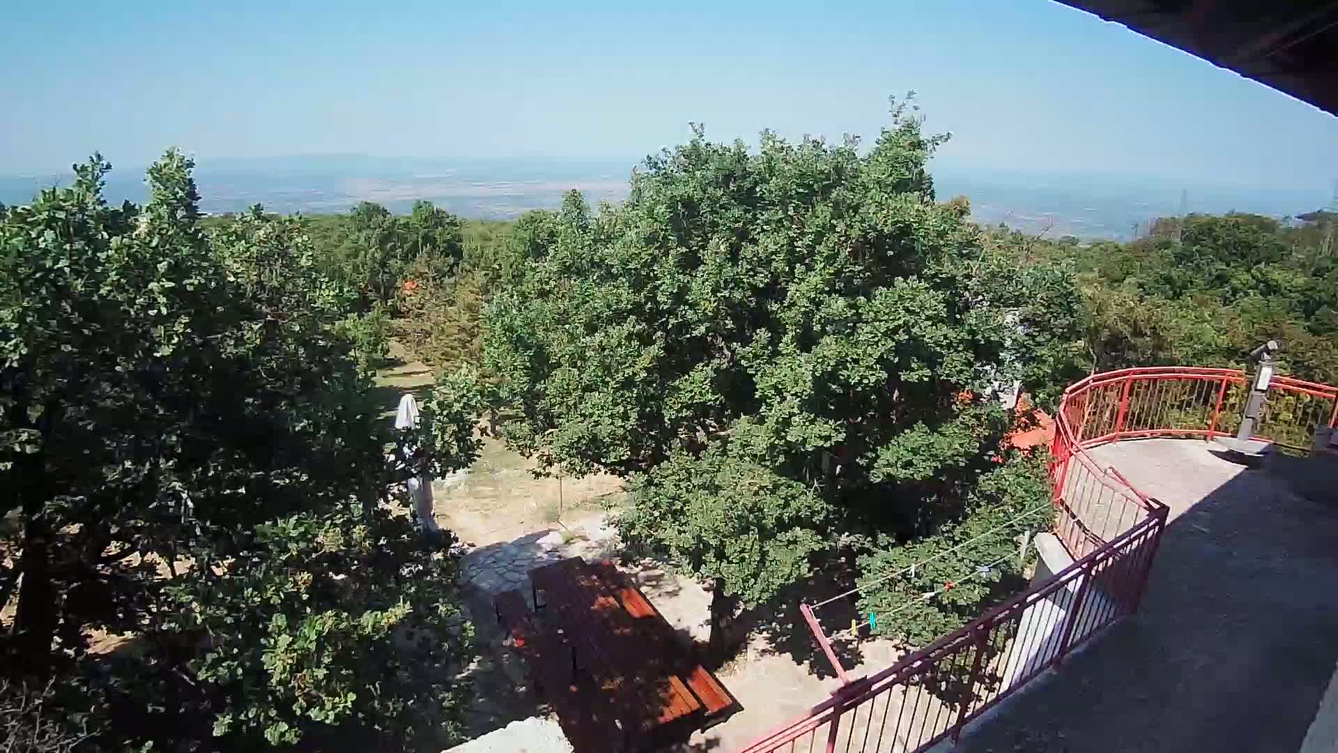 A sunny outdoor scene shows a wooden picnic table beneath leafy green trees, a red railing overlooking a distant valley, and a clothesline with colorful clothing.