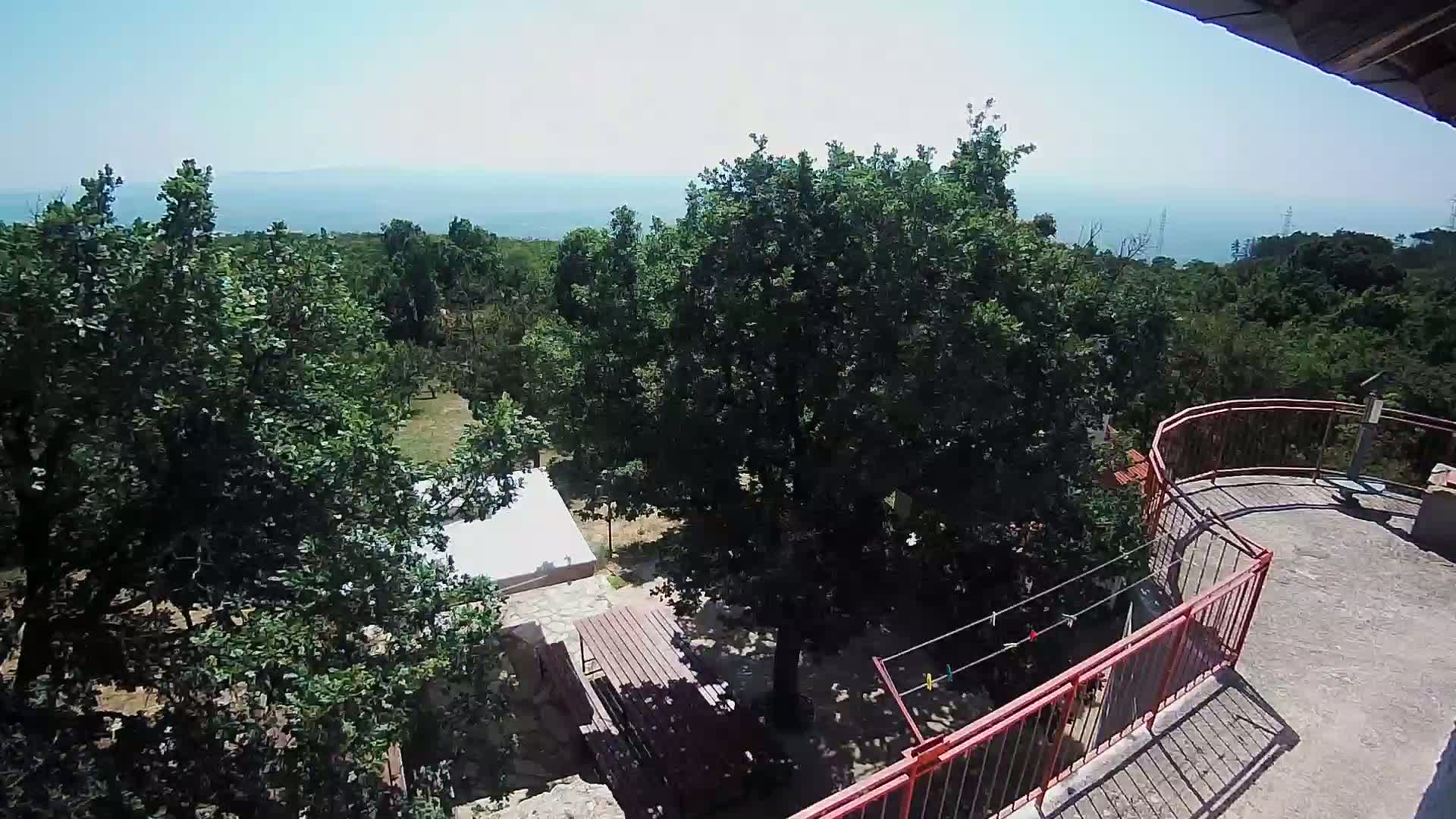 A high-angle view on a sunny day shows a balcony overlooking a wooded area with a patio set, a small building, and a distant ocean.