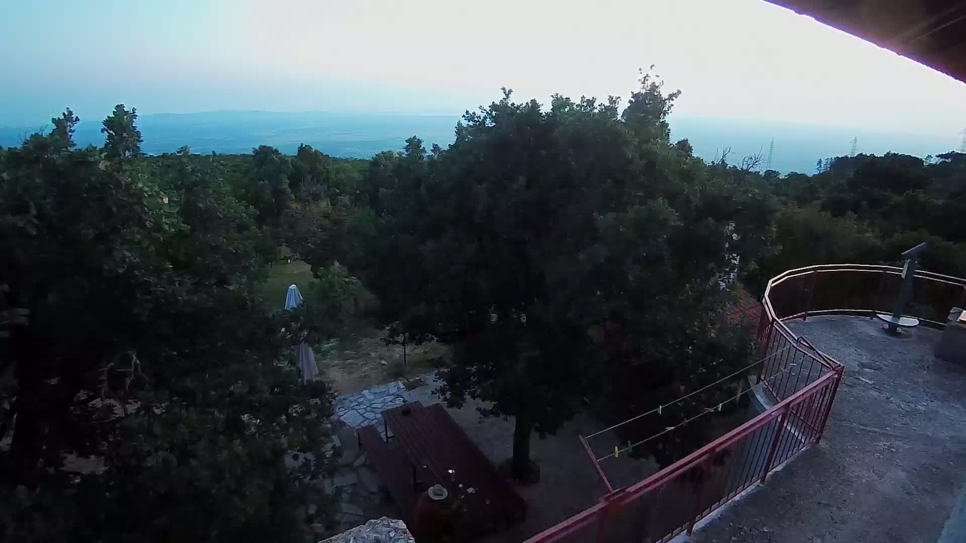 A high-angle view on a sunny day shows a balcony overlooking a wooded area with a patio set, a small building, and a distant ocean.