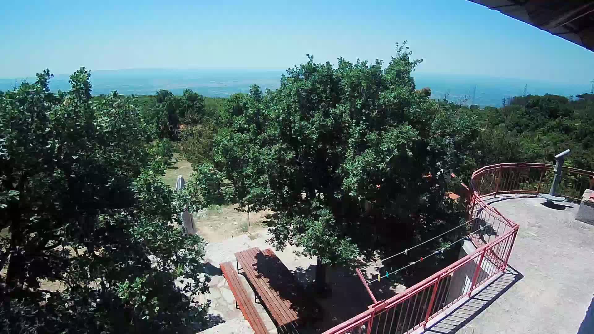 A high-angle view shows a balcony overlooking a sunny, tree-filled yard with picnic tables, a distant sea, and clear blue skies.