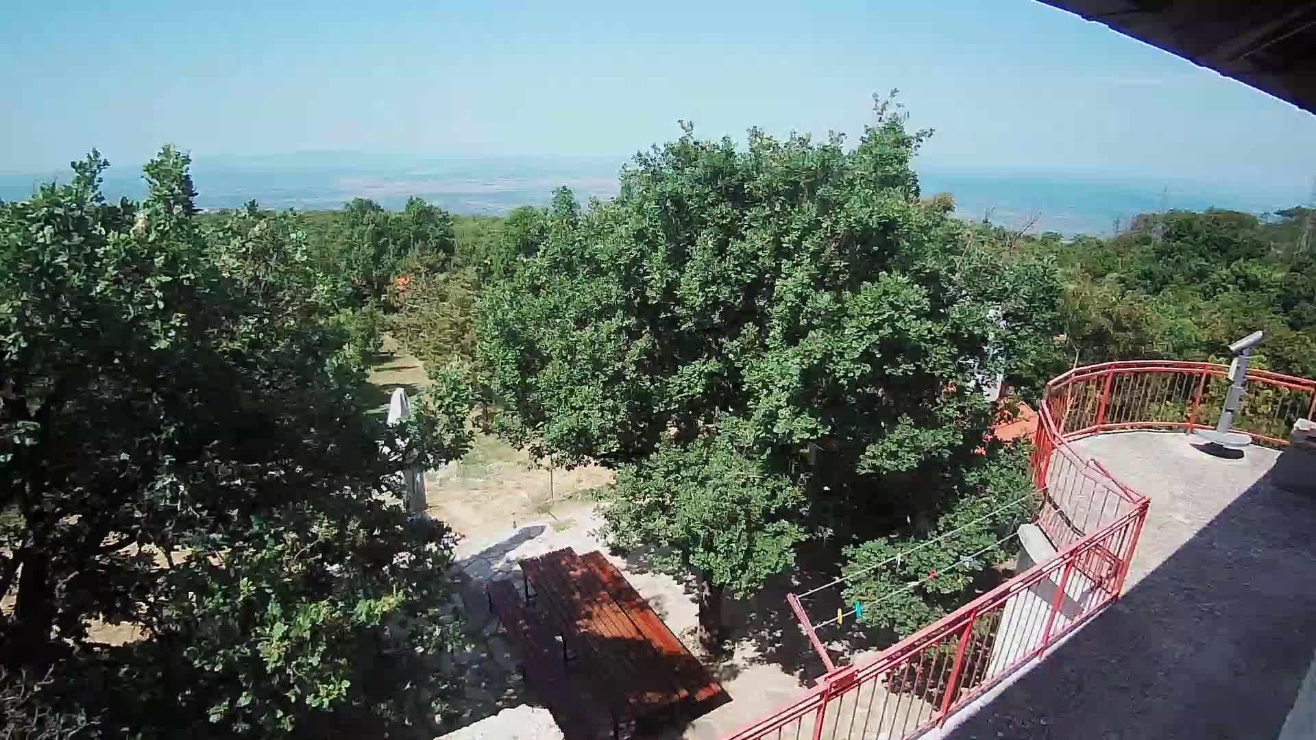 A view from an elevated balcony shows a wooden picnic table below amongst leafy green trees, a red railing surrounding the balcony, and a distant ocean under a clear blue sky.
