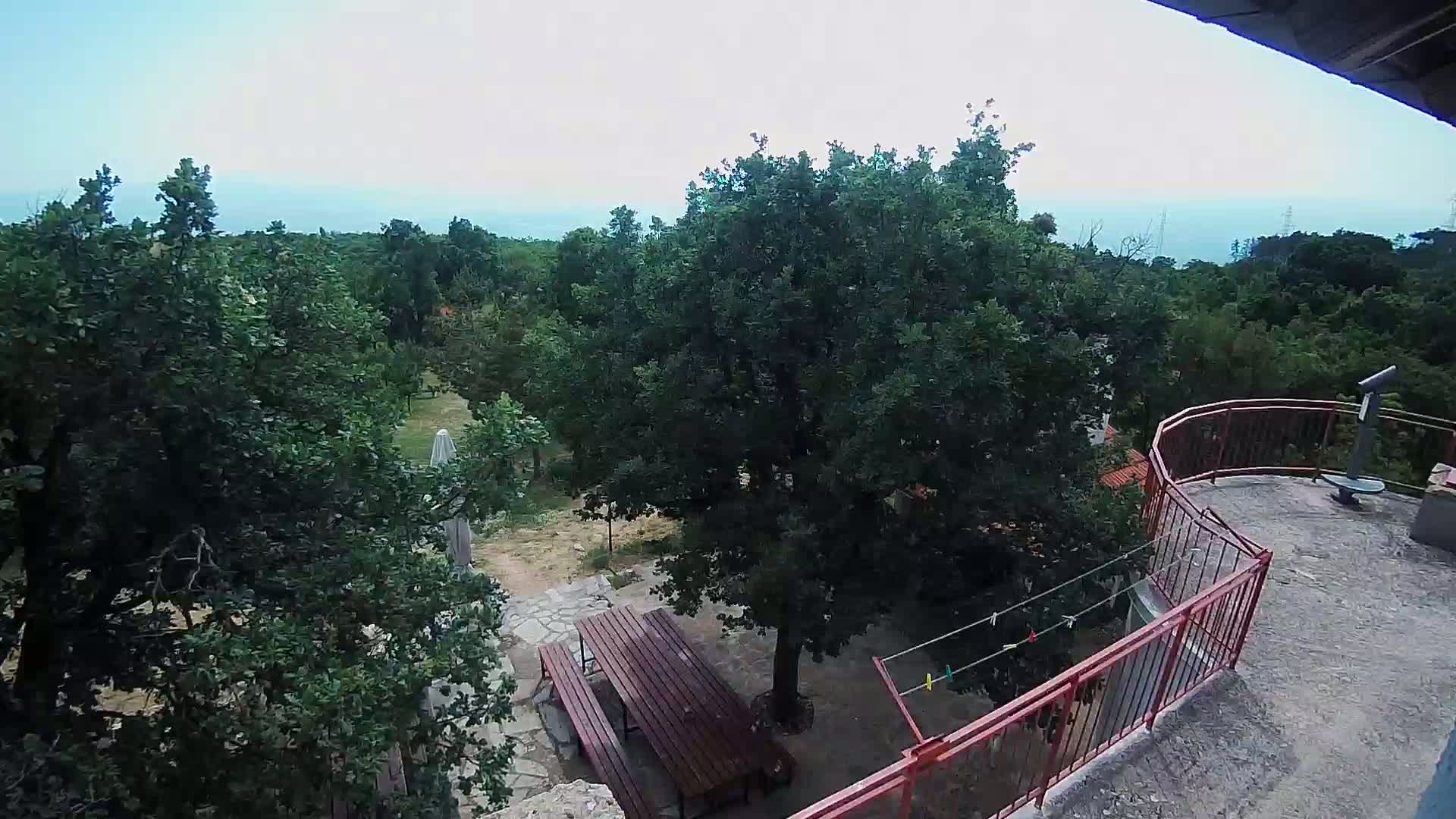 A high-angle view from a balcony shows a partly cloudy day overlooking a wooded area with a picnic table and clothesline below, and a distant ocean in the background.
