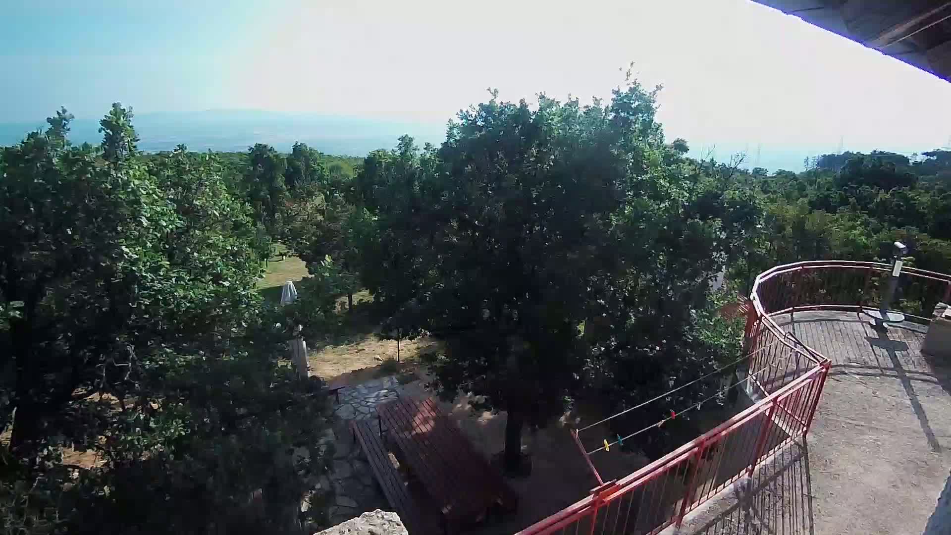 A high-angle view shows a wooden picnic table in a wooded area with a distant ocean view under sunny skies.