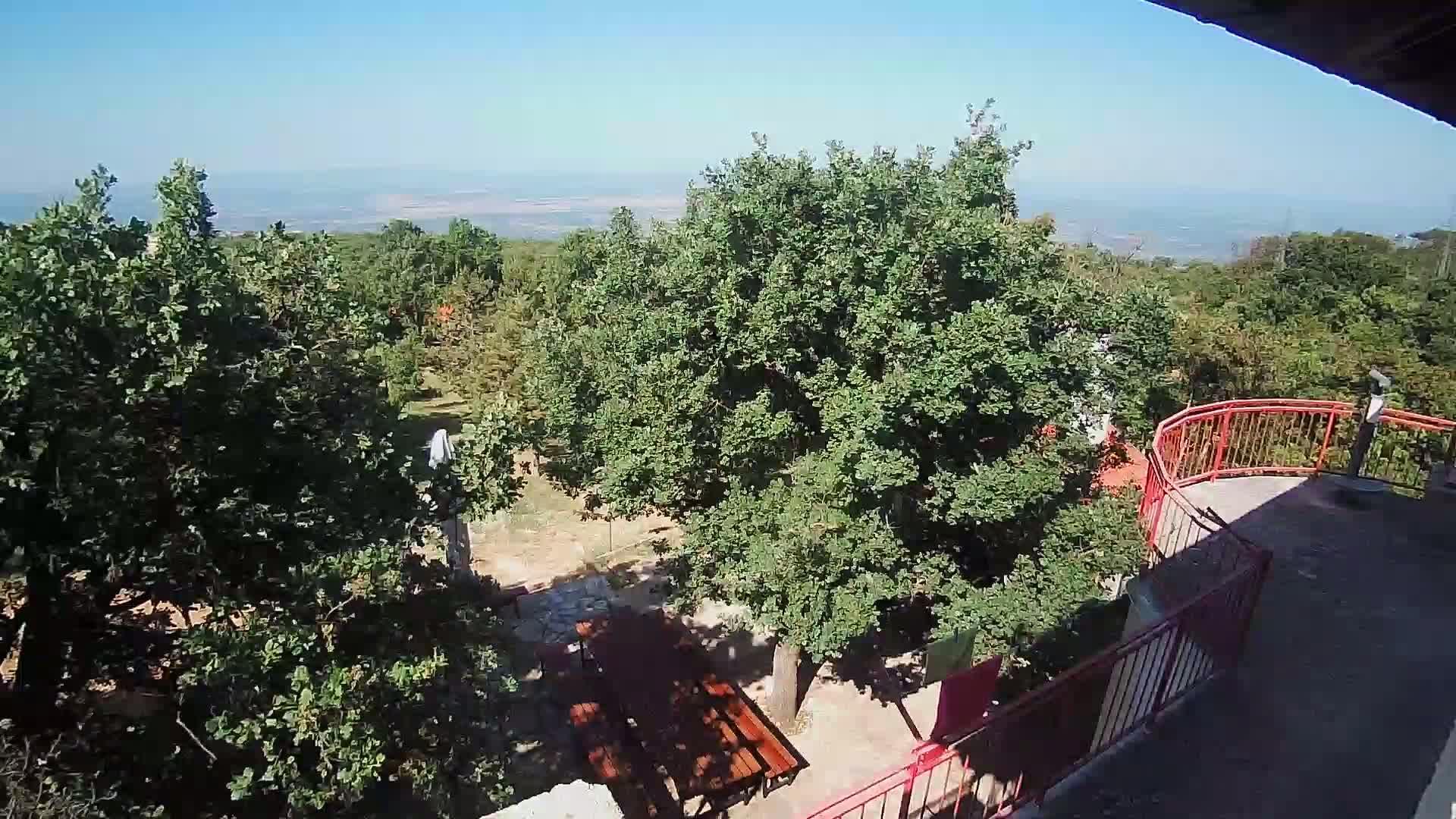 A high-angle view on a sunny day shows a wooden picnic table beneath leafy green trees near a red railing overlooking a distant landscape.