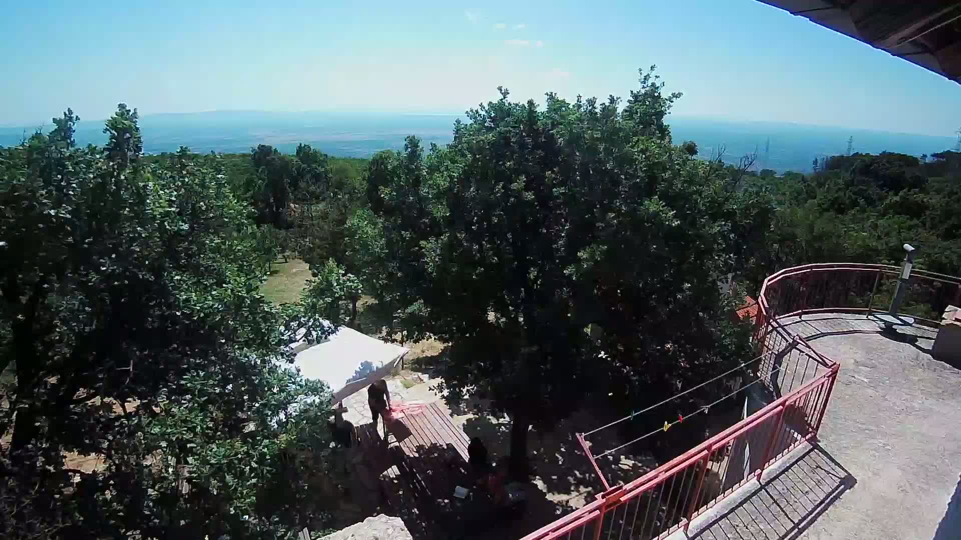 A sunlit outdoor scene shows a balcony overlooking a wooded area with a distant view of a body of water under a clear blue sky.