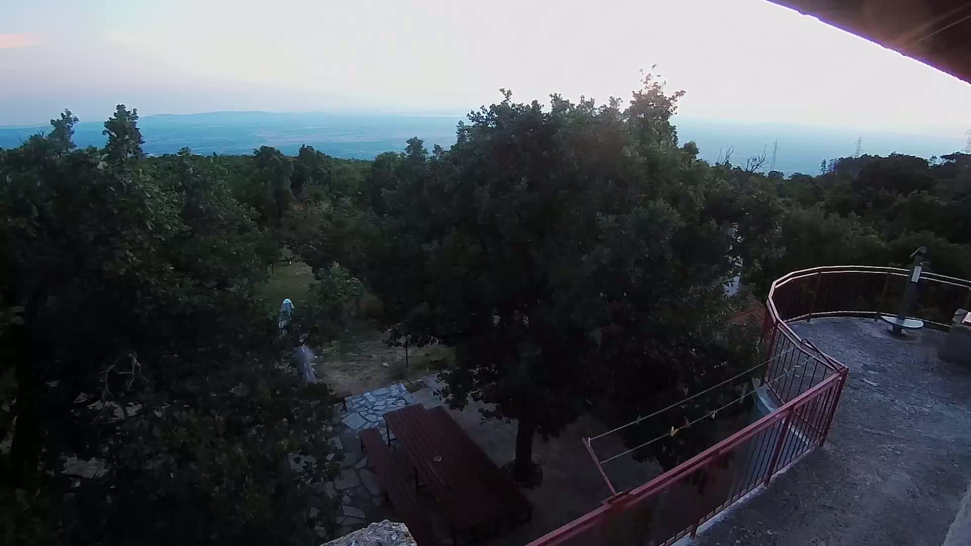 A high-angle view shows a patio with a wooden table and chairs overlooking a forest and a distant body of water under a clear, twilight sky.