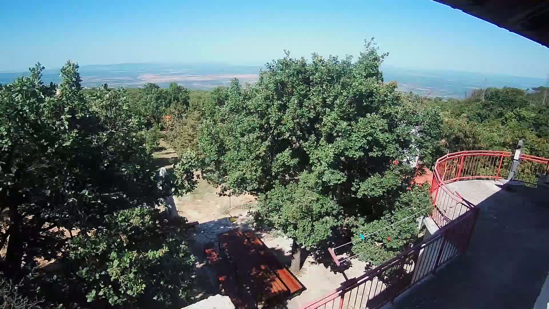 A high-angle view shows a wooden picnic table beneath leafy green trees on a sunny day, overlooking a distant, flat landscape.