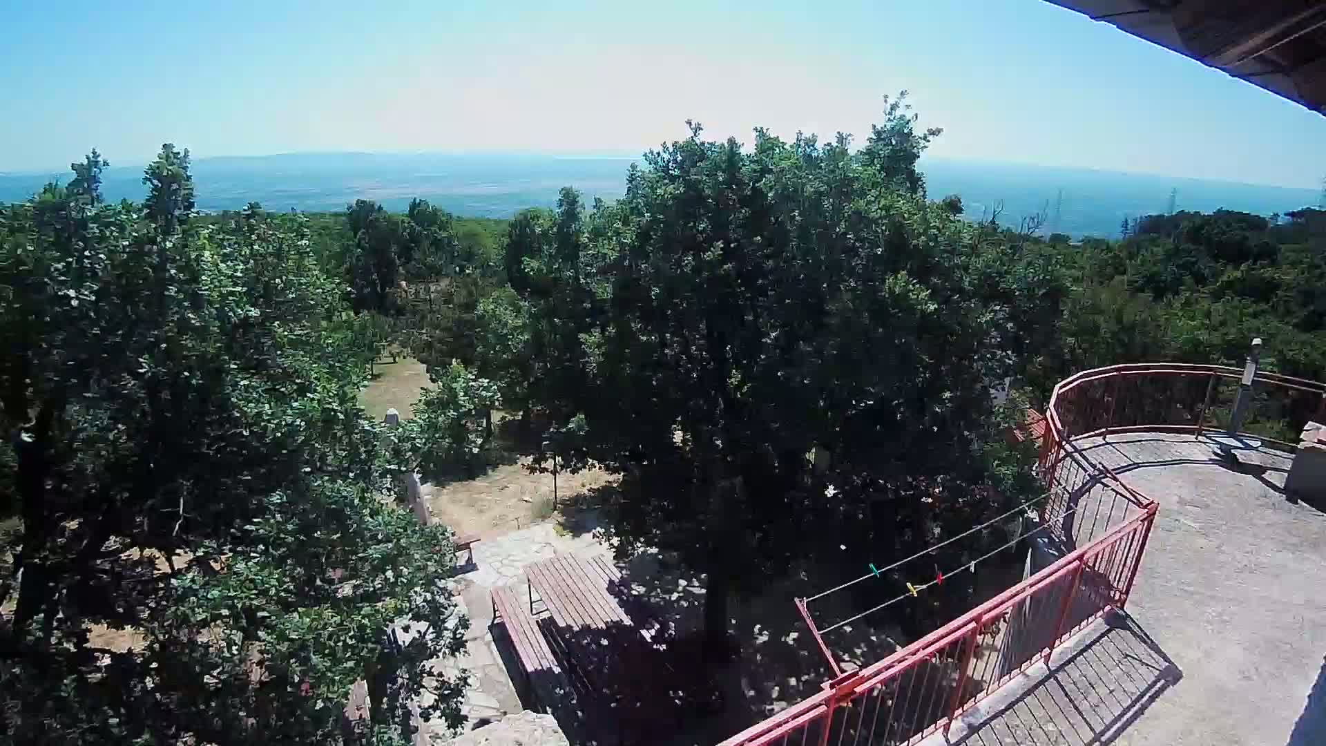 A high-angle view of a stone patio with wooden picnic tables, a red railing, and lush green trees, overlooking a distant body of water under a clear, sunny sky.