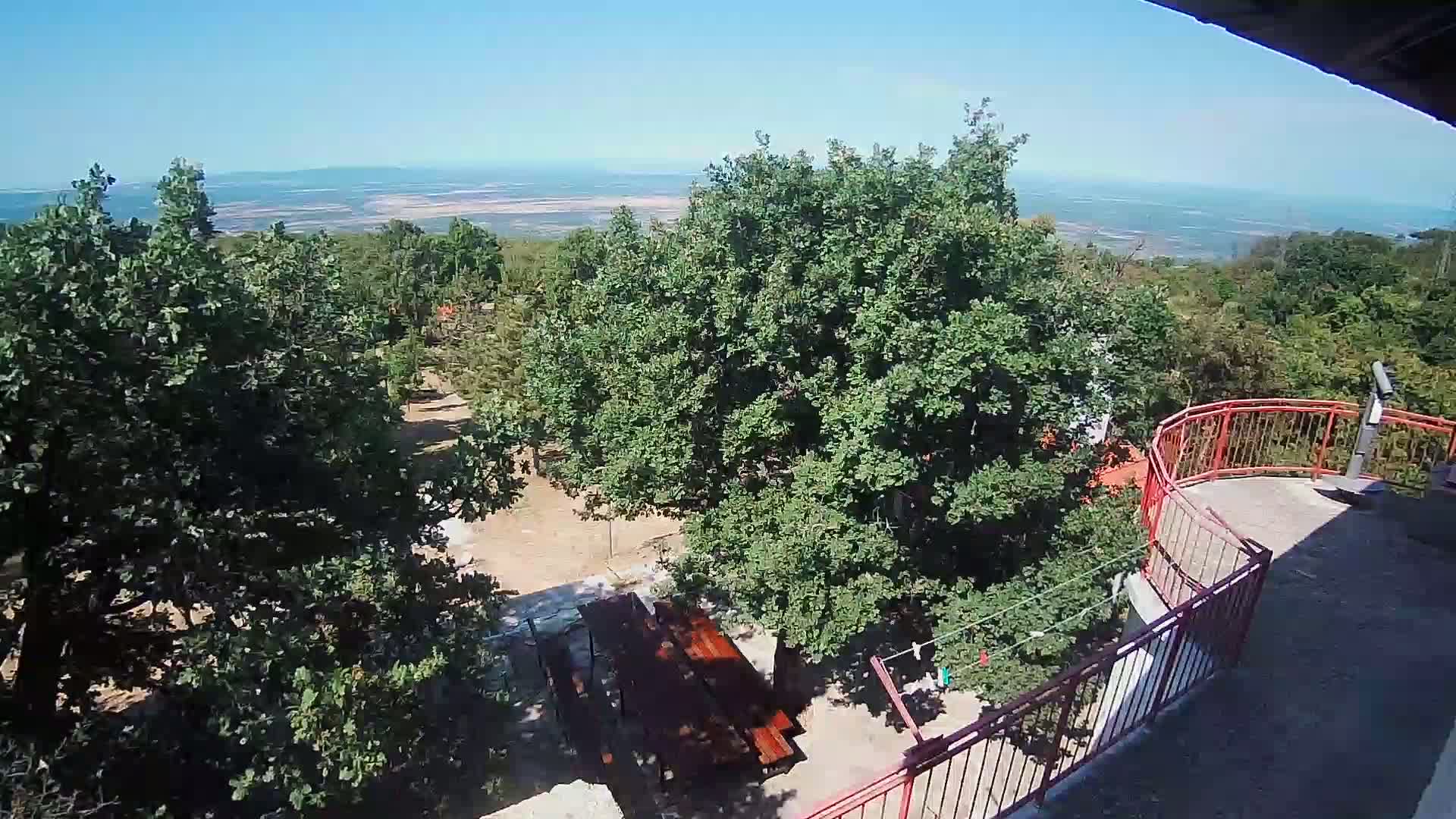 An elevated view shows a picnic table below a red railing on a sunny day overlooking a landscape of green trees and a distant body of water.