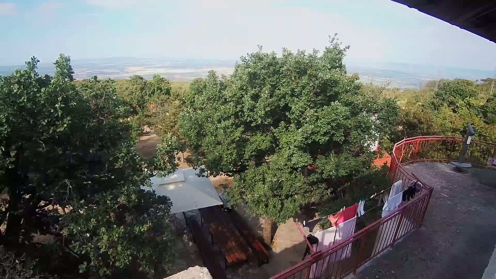 An elevated view shows a tree-filled area with a patio containing a wooden table under a white canopy and clothes drying on a red railing, overlooking a distant body of water under a clear sky.