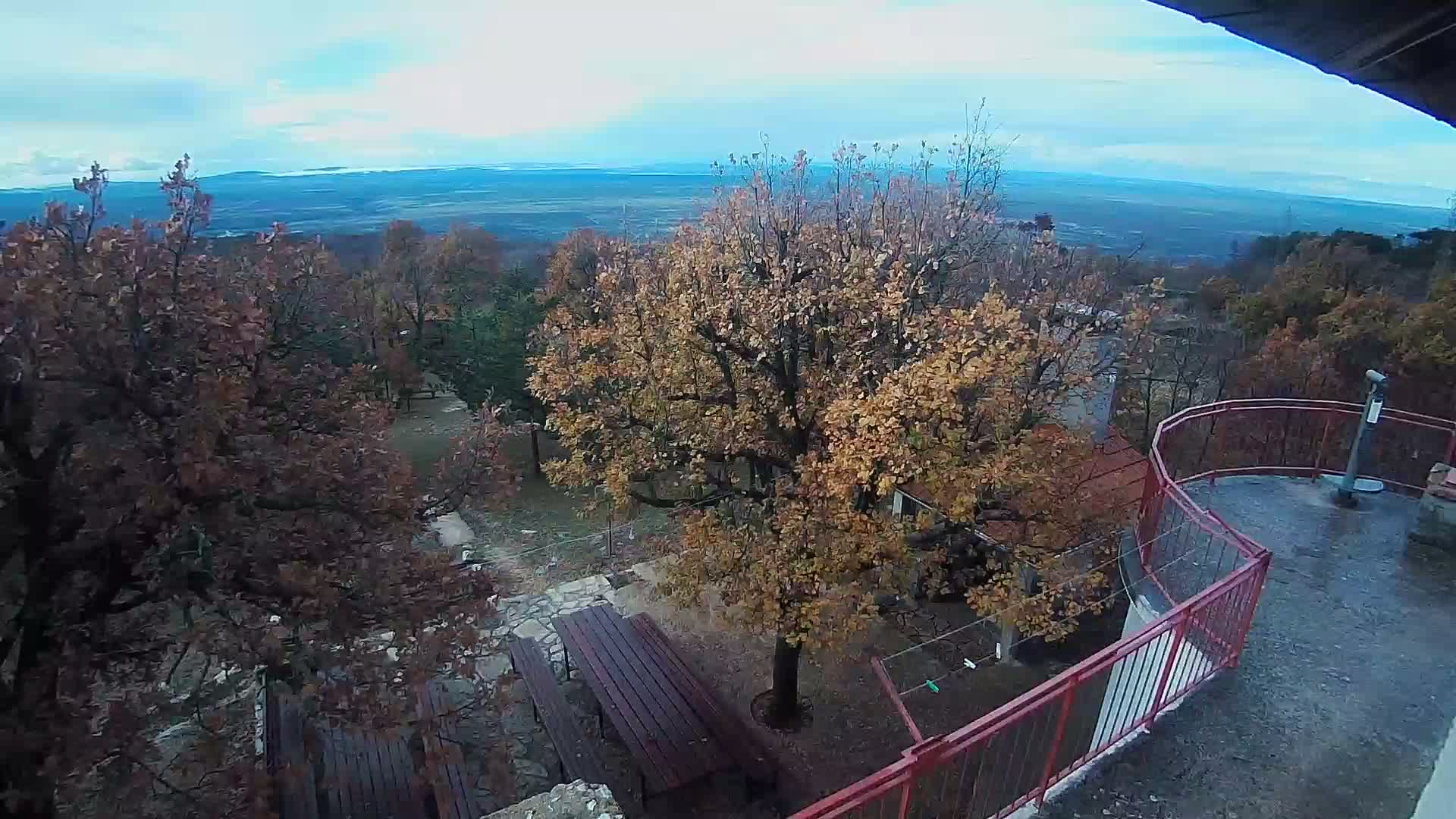 From an elevated perspective, the scene captures autumn trees with brown and yellow foliage, picnic tables, and a viewing platform with a telescope overlooking a distant landscape under a partly cloudy sky.
