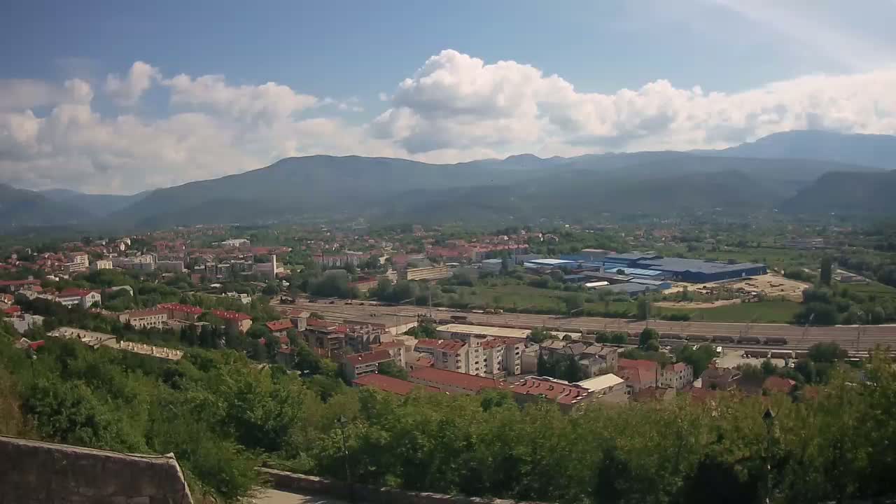 A partly cloudy day reveals a sprawling town nestled in a valley between mountains, with railway lines running through the lower part of the town.