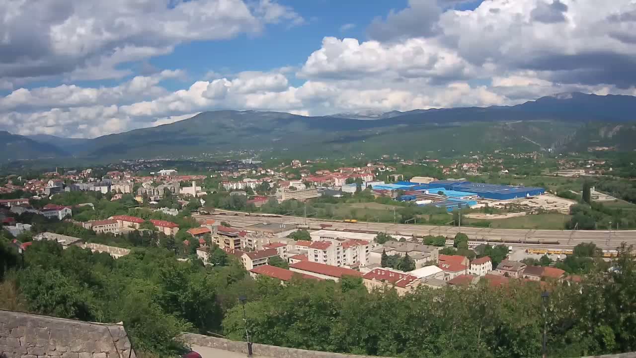 A partly cloudy day reveals a sprawling town nestled in a valley, bordered by mountains, with a train line running through the lower section.