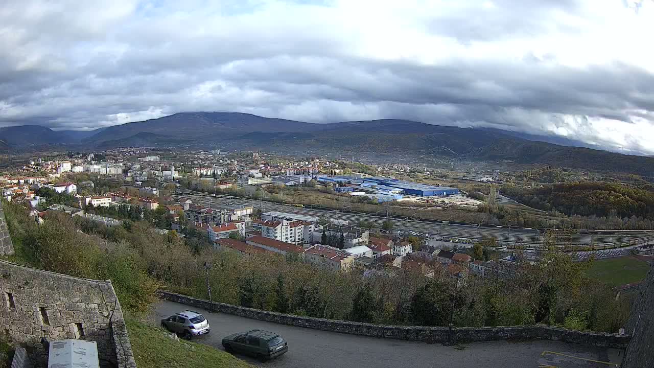 An overcast day reveals a sprawling town nestled in a valley with extensive railway tracks and industrial buildings, all framed by distant mountains under a heavy, cloudy sky, with a winding road and stone walls in the foreground.